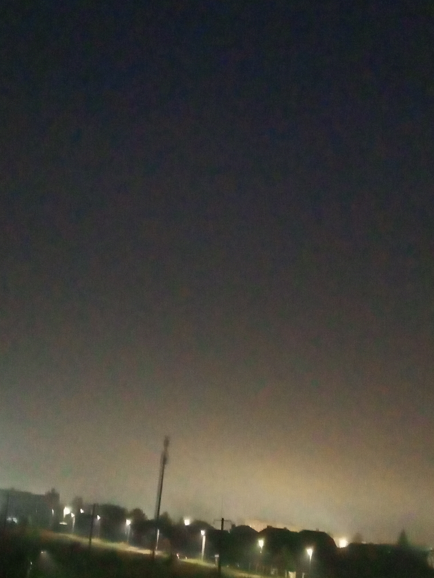 This is a nighttime photograph of an urban or suburban landscape. The sky is dark and appears to be overcast or hazy, with a glow of artificial light emanating from the horizon. In the foreground, there is a dark, grassy area. A tall, slender pole-like structure, possibly a utility pole or antenna, stands prominently on the right side of the frame. Silhouetted against the horizon are the outlines of buildings and trees. Several streetlights are visible, casting pools of light on the ground. The overall impression is of a quiet, dimly lit environment at night. Based on the provided information, this scene is located in the city of Chistopol, Russia.