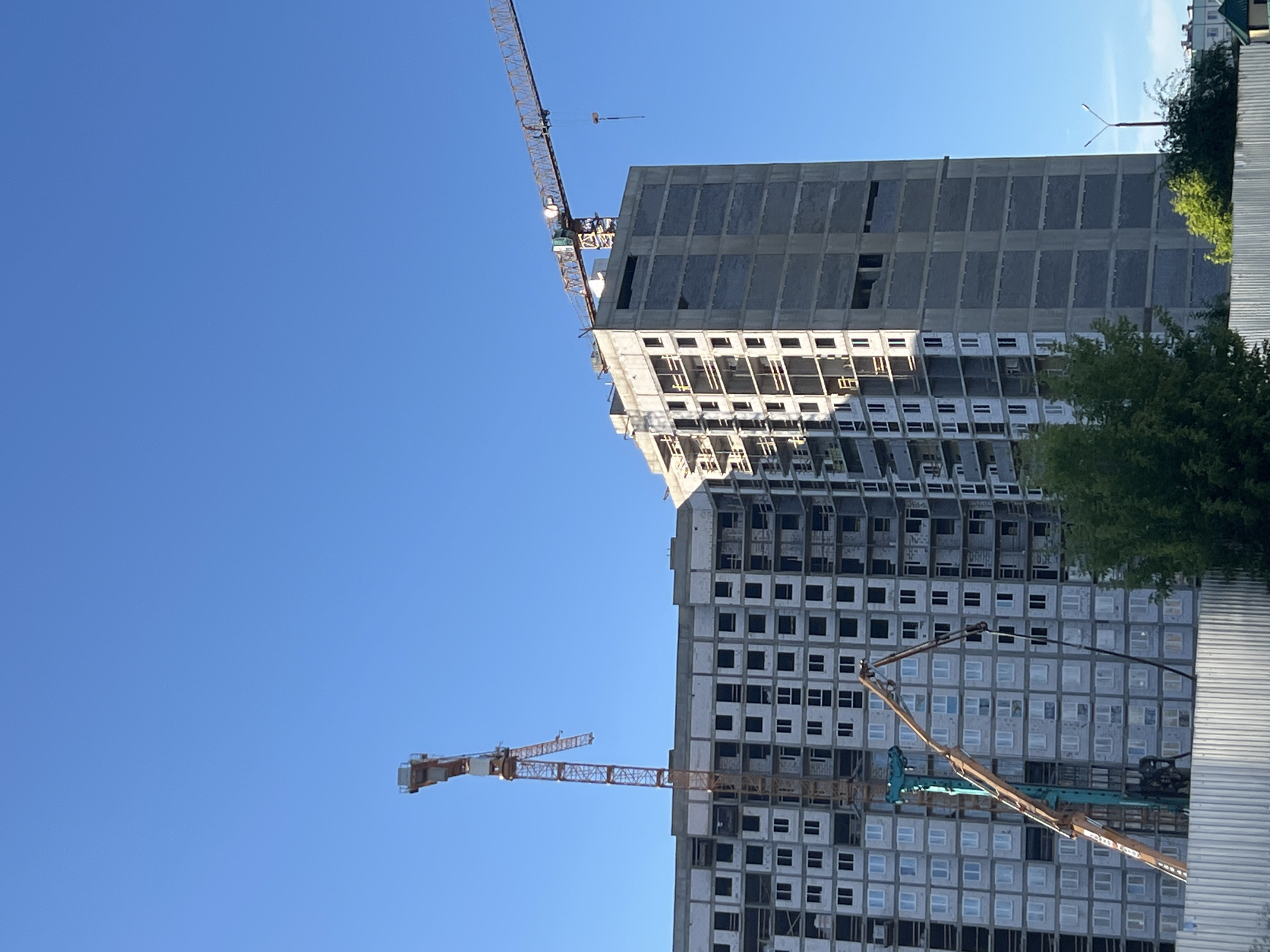 The image shows a construction site with two cranes and unfinished buildings against a clear blue sky. The primary focus is on a tall, multi-story building under construction. The building's facade is made of concrete panels, and many of the window openings are empty or covered with temporary materials. In the foreground, a large construction crane with a yellow jib extends across the frame. Another, smaller crane is visible further in the distance.
The scene suggests a typical day of construction work. The bright blue sky indicates fair weather and likely daytime. The urban setting is implied by the scale of the buildings and the presence of construction equipment. No people are clearly visible, but their absence doesn't detract from the depiction of an ongoing construction project. The overall impression is one of industrial activity and development in an urban environment. There is no visible text in the image. The context provided states the location is Minsk, Belarus.