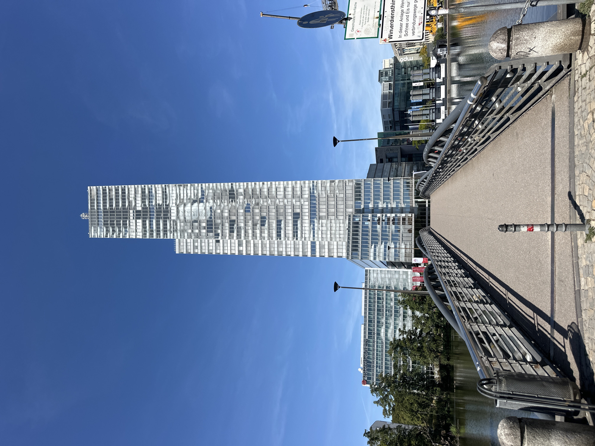 The image shows a modern pedestrian bridge leading towards a tall, glass-facade skyscraper in the city of Cologne, Germany. The skyscraper is a prominent feature, with its reflective windows mirroring the clear blue sky. To the left of the skyscraper, there are other modern buildings and lush green trees surrounding a body of water, likely a canal or river. The bridge itself is made of metal with a paved walkway. Signs are visible on the right side of the image, one indicating "Geschützte Freifläche" (Protected Open Space) with rules about not disturbing wildlife, and another sign related to winter services ("Winterdiensthinweis") stating that snow and ice removal is only for access routes. The overall scene depicts an urban environment with a blend of modern architecture and natural elements, under a bright, sunny sky.