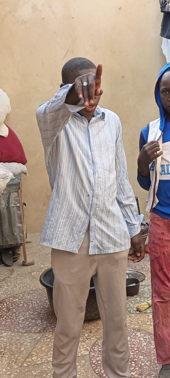 The image features two young individuals in what appears to be an outdoor or semi-outdoor setting in Kaura Goje, Nigeria, under bright daylight.
The central figure is a young person with dark skin and short, dark hair, wearing a light blue and white striped long-sleeved button-up shirt and light brown/khaki trousers. This individual is making a gesture with their right hand, with the index finger pointing upwards, partially obscuring their face. A silver-colored ring with a rectangular setting is visible on their right ring finger. Their mouth is slightly open, possibly indicating speech or a reaction. The light brown trousers have several noticeable damp spots or stains.
To the right, partially visible, is another young person with dark skin, wearing a blue hooded top with white accents and red or pink trousers. Only a portion of their face and upper body is visible. They are looking towards the camera, with their right hand resting near their chest. Their red trousers also display damp spots.
The background consists of a plain, light brown wall. To the far left, a white pillow or plush toy and a red cushion are stacked on a low structure. A corrugated metal object is also partially seen on the left. The ground is covered in a multi-colored mosaic tile pattern, incorporating shades of beige, brown, and white. A black plastic basin sits on the ground between the two people. Closer to the right-hand subject, a small dark pot, a metal tray, and a traditional straw broom are visible on the ground.
The overall scene suggests an informal, possibly domestic or communal environment. No specific activity beyond standing and gesturing is explicitly taking place, though the damp clothing on both individuals might imply recent interaction with water or liquids. There is no clearly legible text visible anywhere in the image.