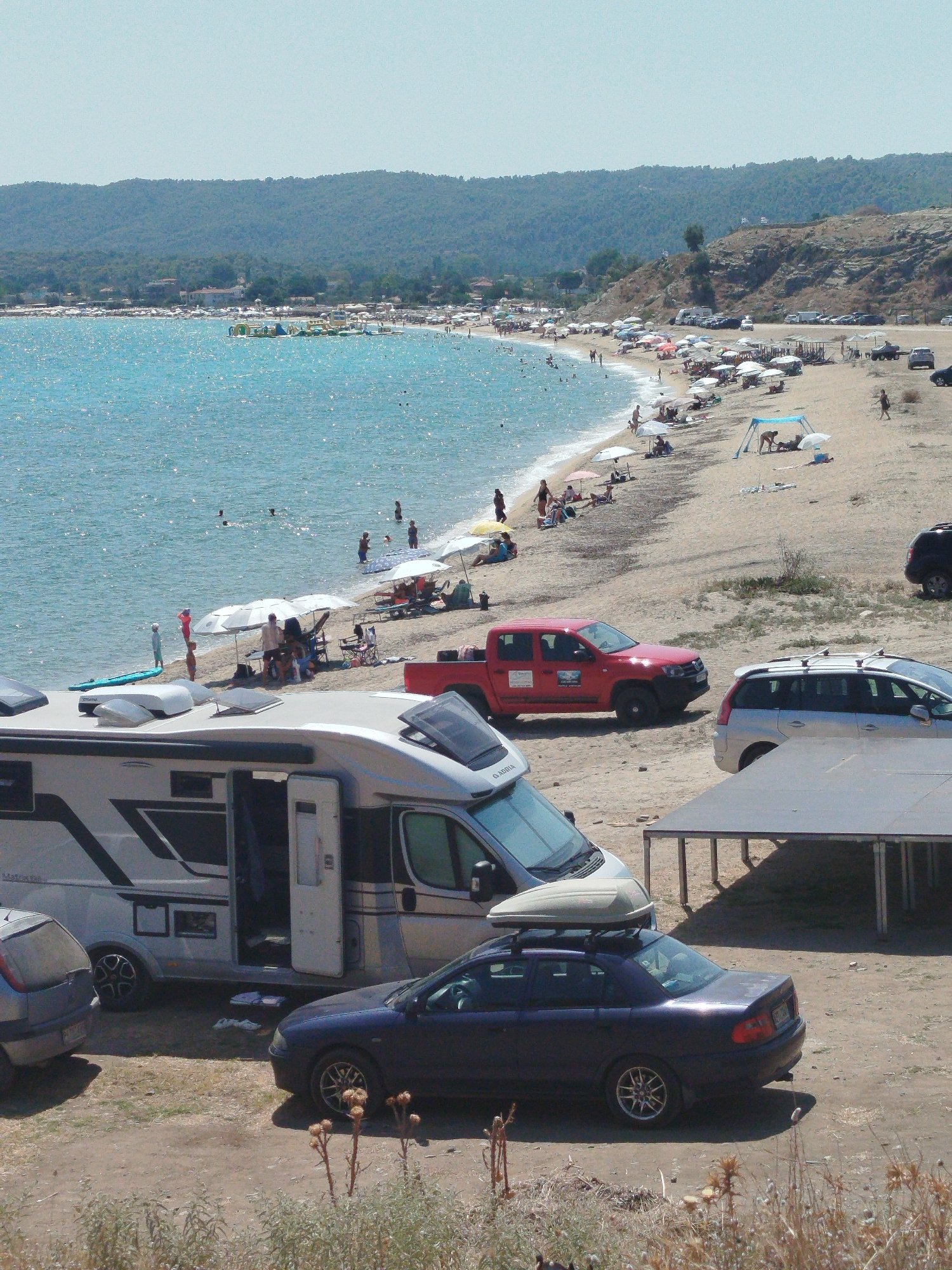 A high-angle medium shot captures a sun-drenched beach in Sithonia Municipal Unit, Greece, populated with numerous beachgoers and vehicles. The turquoise waters of the Aegean Sea lap gently onto a sandy shore lined with rows of beach umbrellas and lounge chairs. In the foreground, a silver RV and a dark blue sedan are parked on the dusty ground. The RV, with its door open, faces left, while the sedan is parked to its right. Further back, a red pickup truck is parked near the beach, and a silver SUV is seen closer to the water. People are scattered across the beach, some swimming in the sea, others relaxing under umbrellas, and a few walking along the shoreline. The background features a gentle, tree-covered hill sloping down towards the coast. The scene conveys a relaxed, holiday atmosphere.