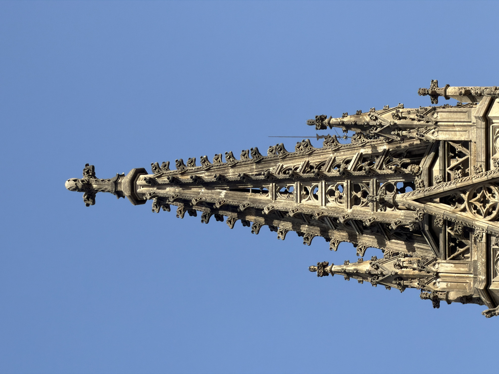 The image shows the ornate spires of the Barcelona Cathedral against a clear blue sky. The cathedral's Gothic architecture is characterized by intricate stone carvings, pointed arches, and flying buttresses. The spires are topped with crosses and decorated with various gargoyles and decorative elements. The lighting suggests it is daytime, with strong sunlight casting shadows on the stone. No people are visible in the image. The setting is clearly an urban environment, specifically the city of Barcelona, Spain, given the architectural style and the mention of the Barcelona Cathedral.