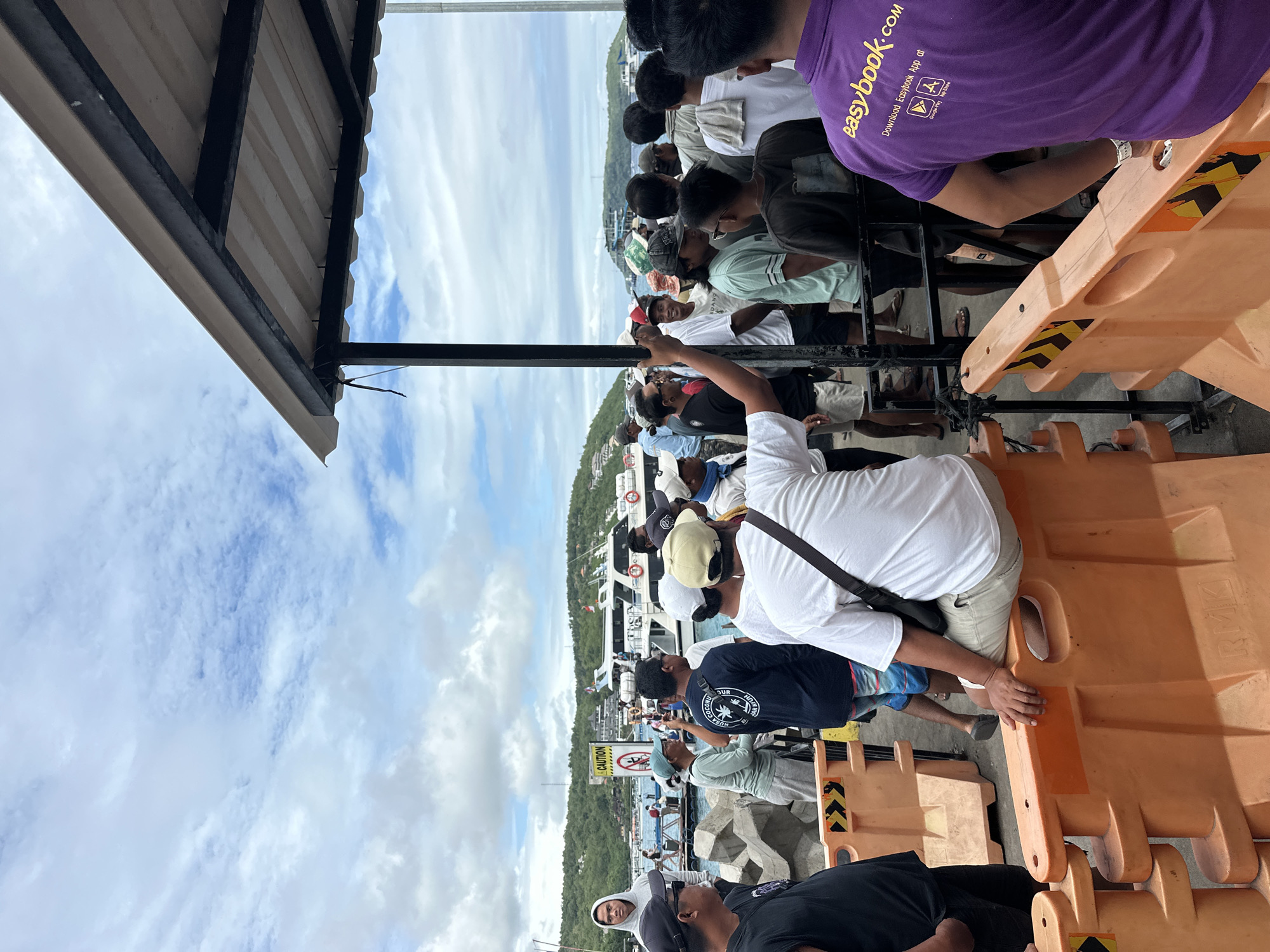 People are lined up at a waterfront embarkation point at Nusa Penida, Indonesia. They are positioned behind orange plastic crowd control barriers on a pier, and there is a roof to the left. The crowd looks toward a boat.