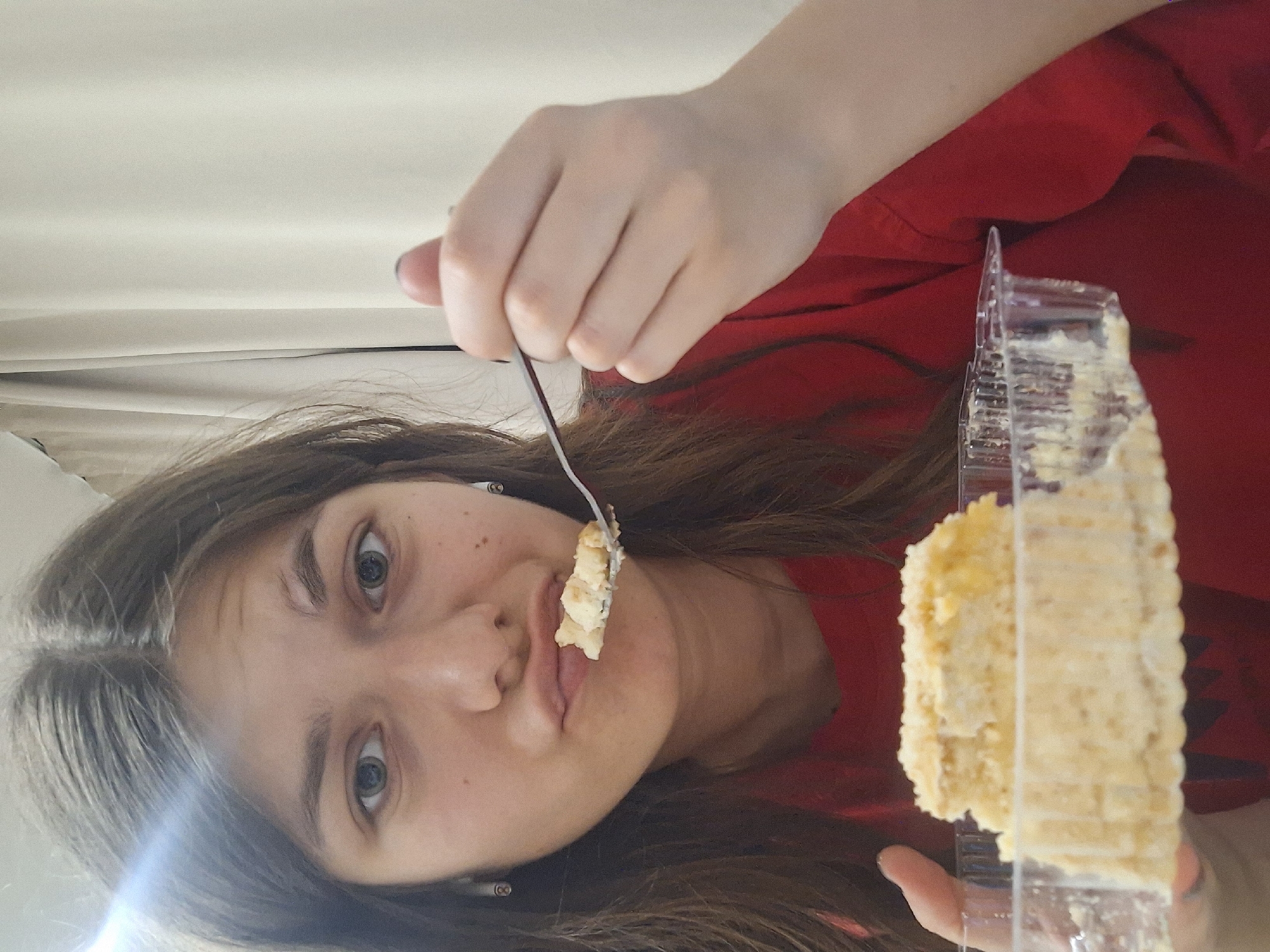 A woman is holding a fork with a piece of cake near her mouth and holding the remainder of the cake in a clear plastic container. The woman has light skin, brown hair, and is wearing a red shirt. The background appears to be a white or light-colored wall. It is not possible to determine the location in the city of Panagyurishte, Bulgaria from the image.