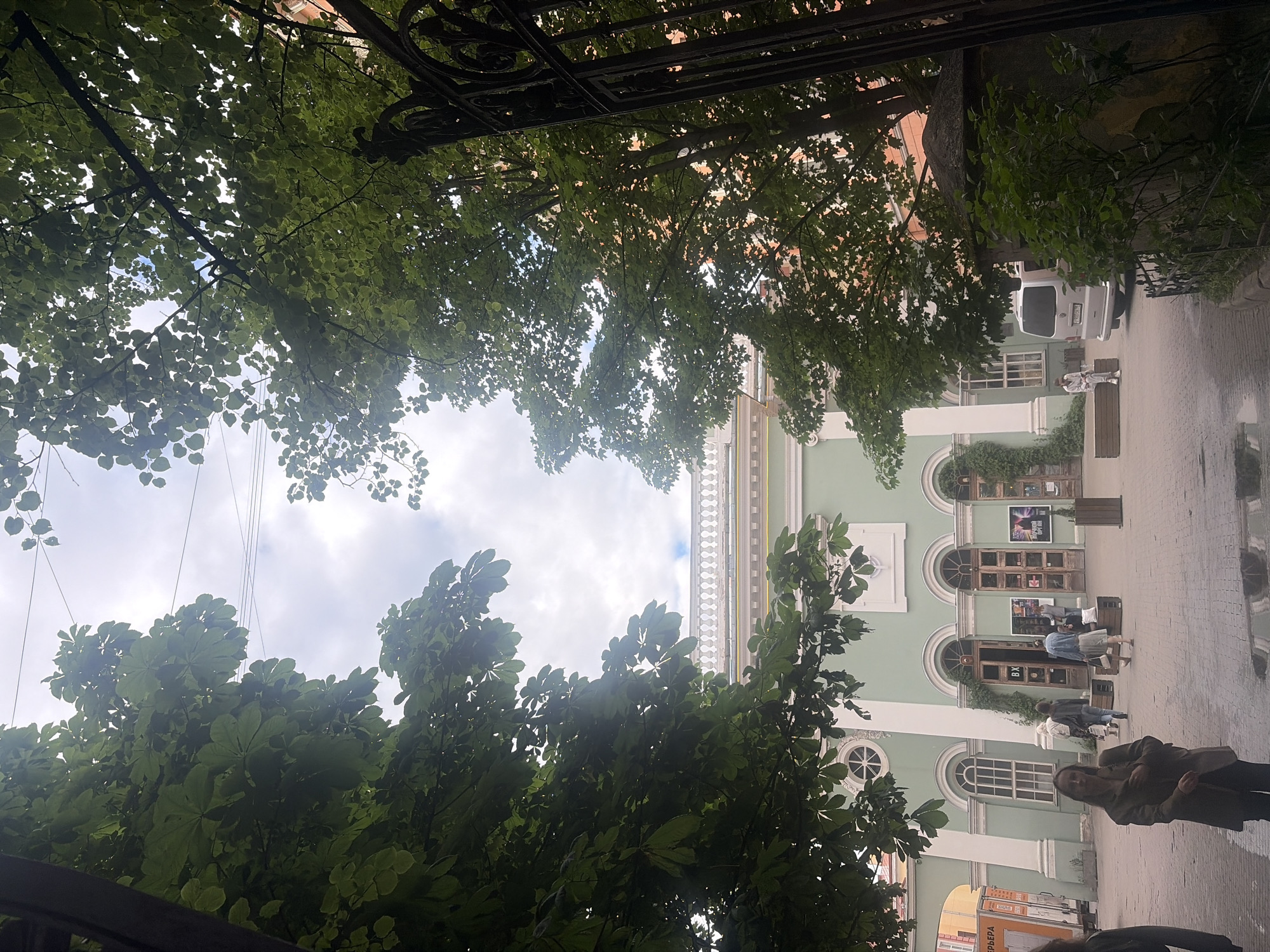 A paved courtyard is depicted, with a pale green building featuring arched doorways and windows in the background. Lush green trees with dense foliage frame the top and sides of the image. Several people are visible in the courtyard, some walking towards the building, others standing or sitting. A light-colored van is parked to the right of the building. The sky is cloudy. The building's architecture, particularly the decorative cornice, suggests it is a cultural institution, possibly a theater or museum. Based on the architectural style and the presence of what appears to be Russian signage on posters near the entrances, the location is likely Saint Petersburg, Russia.