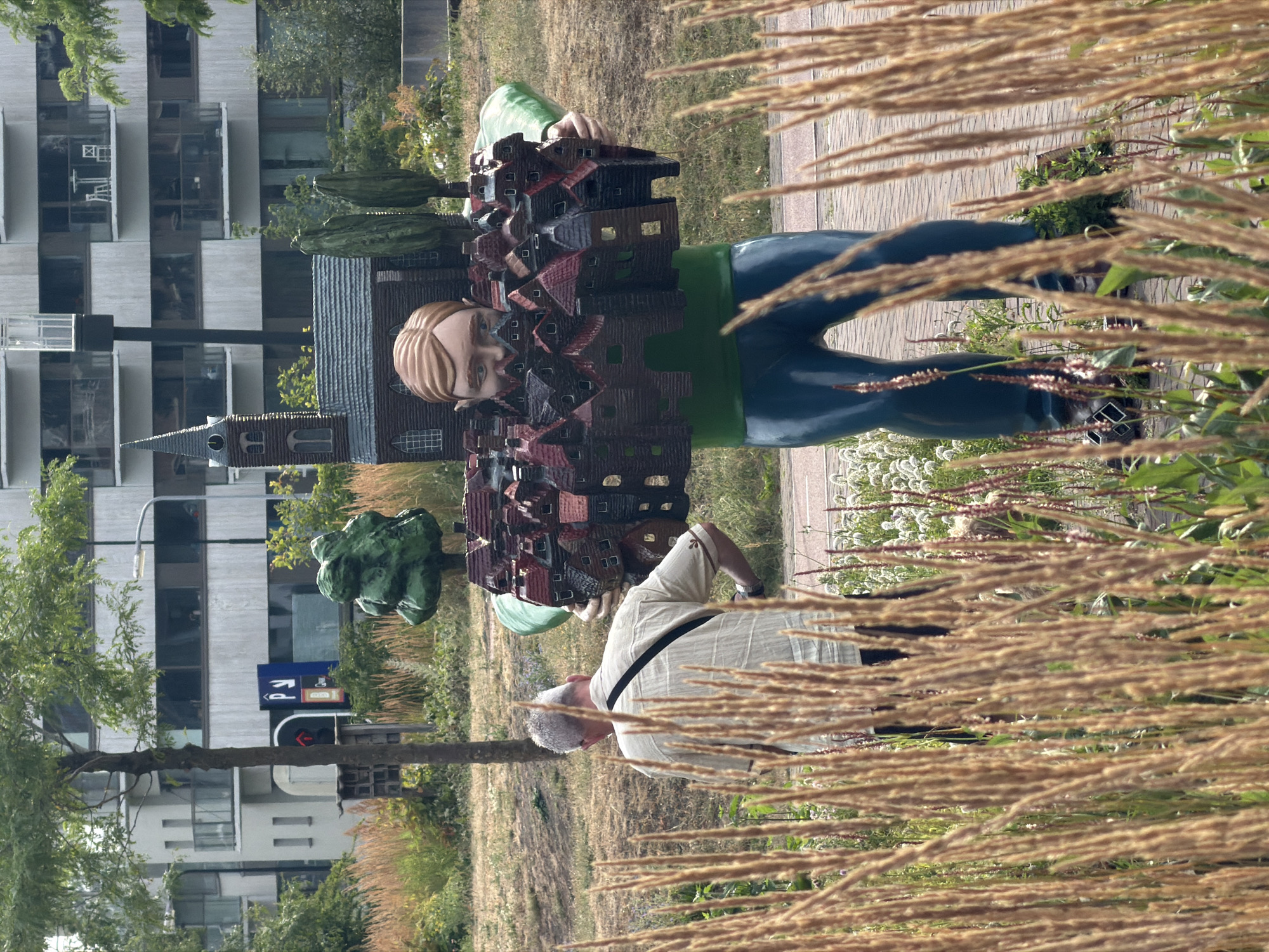 A life-sized statue of a figure is depicted holding a cluster of miniature houses and a church around its torso, in the city of Rijswijk, Netherlands. The statue has light brown hair, a light green shirt, and dark blue pants, with its face obscured by the houses. A man with grey hair wearing a light-colored short-sleeved shirt and black pants stands to the left of the statue, facing away from the camera. The statue and the man are situated in an outdoor environment with tall, dry grass in the foreground and a blurred background featuring a building, trees, and a street. The statue is positioned on what appears to be a paved surface with some greenery around its base.