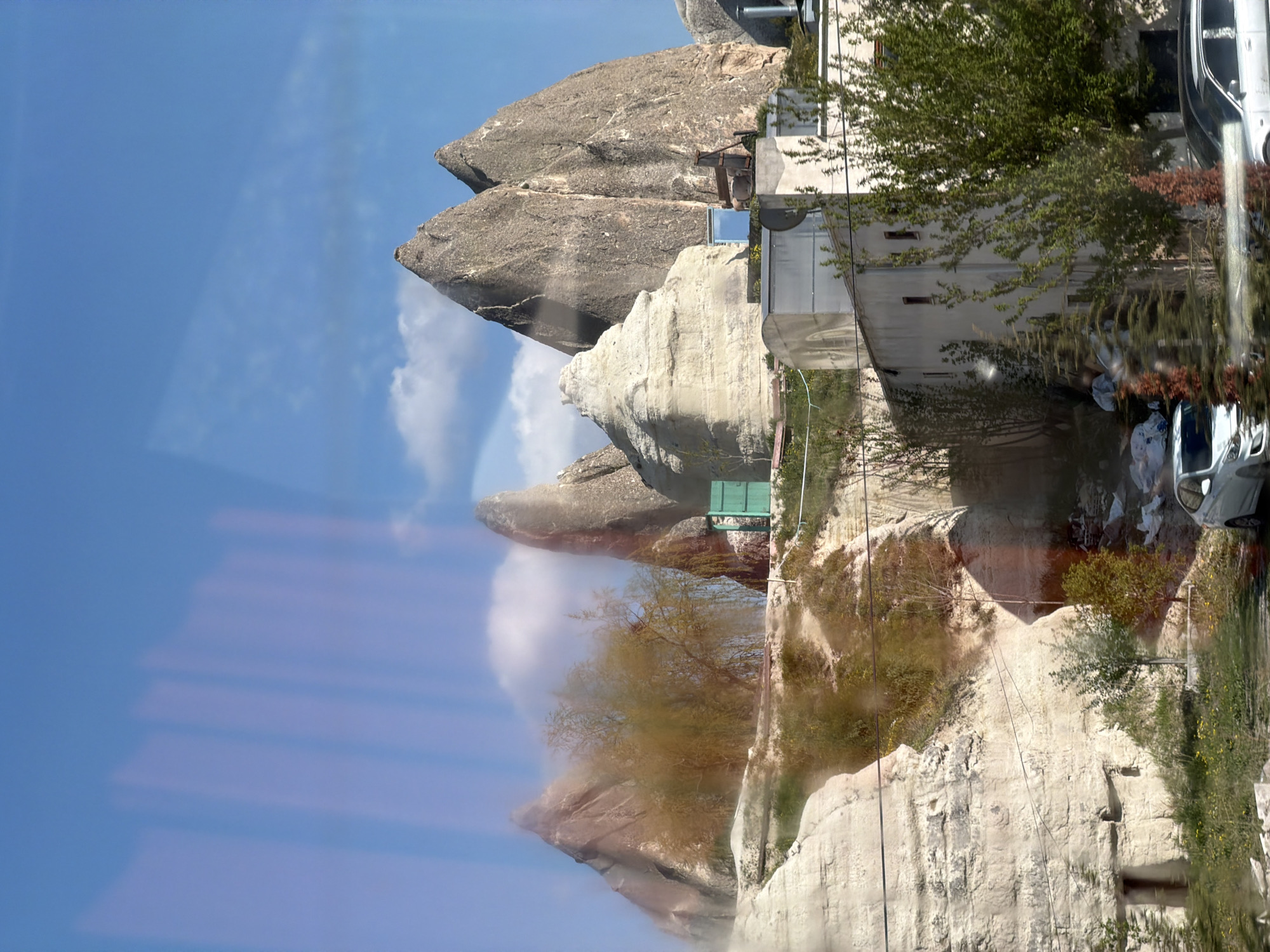 This daytime image showcases the unique landscape of Göreme, Cappadocia, Turkey, characterized by its distinctive "fairy chimney" rock formations. The photograph appears to have been taken through a window, as evidenced by prominent reflections of the bright blue sky and some blurred internal elements visible on the left and lower parts of the frame.
The scene is dominated by tall, conical and pillar-shaped rock formations, ranging in color from light brown to grey, indicative of their volcanic tuff composition. A clear blue sky with a few scattered white clouds provides a serene backdrop, suggesting fair and sunny weather, likely midday or early afternoon.
Human structures are intricately integrated into this natural environment. Several buildings, possibly hotels or homes, with concrete walls and flat roofs, are visible on the right side of the image, nestled directly against or carved into the rock formations. Green trees and shrubs are interspersed among the rocks and buildings, adding organic texture to the scene. Utility lines or wires traverse parts of the landscape.
On the far right, parts of a light-colored car are visible, parked or moving. Another vehicle is partially seen further down. A distinct green bench or platform is situated on one of the higher rock formations. No people are visible, and there is no discernible text in the image. The overall impression is one of harmonious coexistence between a remarkable natural landscape and human settlement.