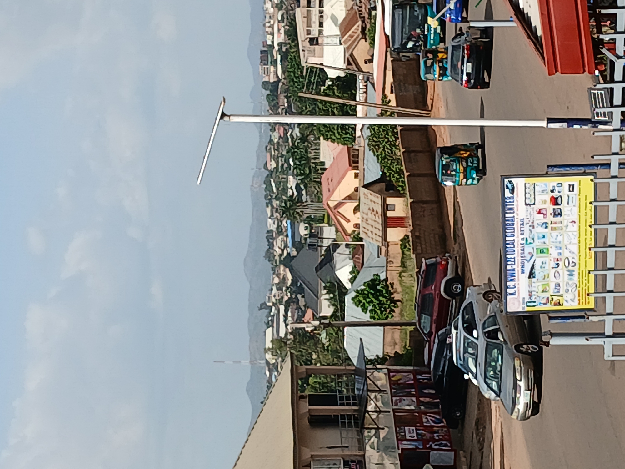 This is a medium-distance, eye-level shot of a street in Jos, Nigeria, on a clear, sunny day. The scene captures a typical urban environment with a mix of residential and commercial buildings, a busy road with vehicles, and distant hills.
In the foreground, a paved road runs across the image. A silver sedan is driving away from the viewer, and to its left, a darker sedan is parked. Further down the road, a red SUV is parked. To the right of the road, a blue and white bus is visible. A yellow and green auto-rickshaw is also present on the road. A white lamppost with a solar panel on top stands prominently on the right side of the road.
In the mid-ground, various buildings of different architectural styles are visible. Some are single-story structures, while others have multiple levels. Roofs are predominantly red or grey. Green trees and foliage are scattered throughout the area.
In the background, a hazy view of distant hills under a pale blue sky with scattered white clouds completes the scene. The overall impression is one of a moderately populated urban area with ongoing daily activities.