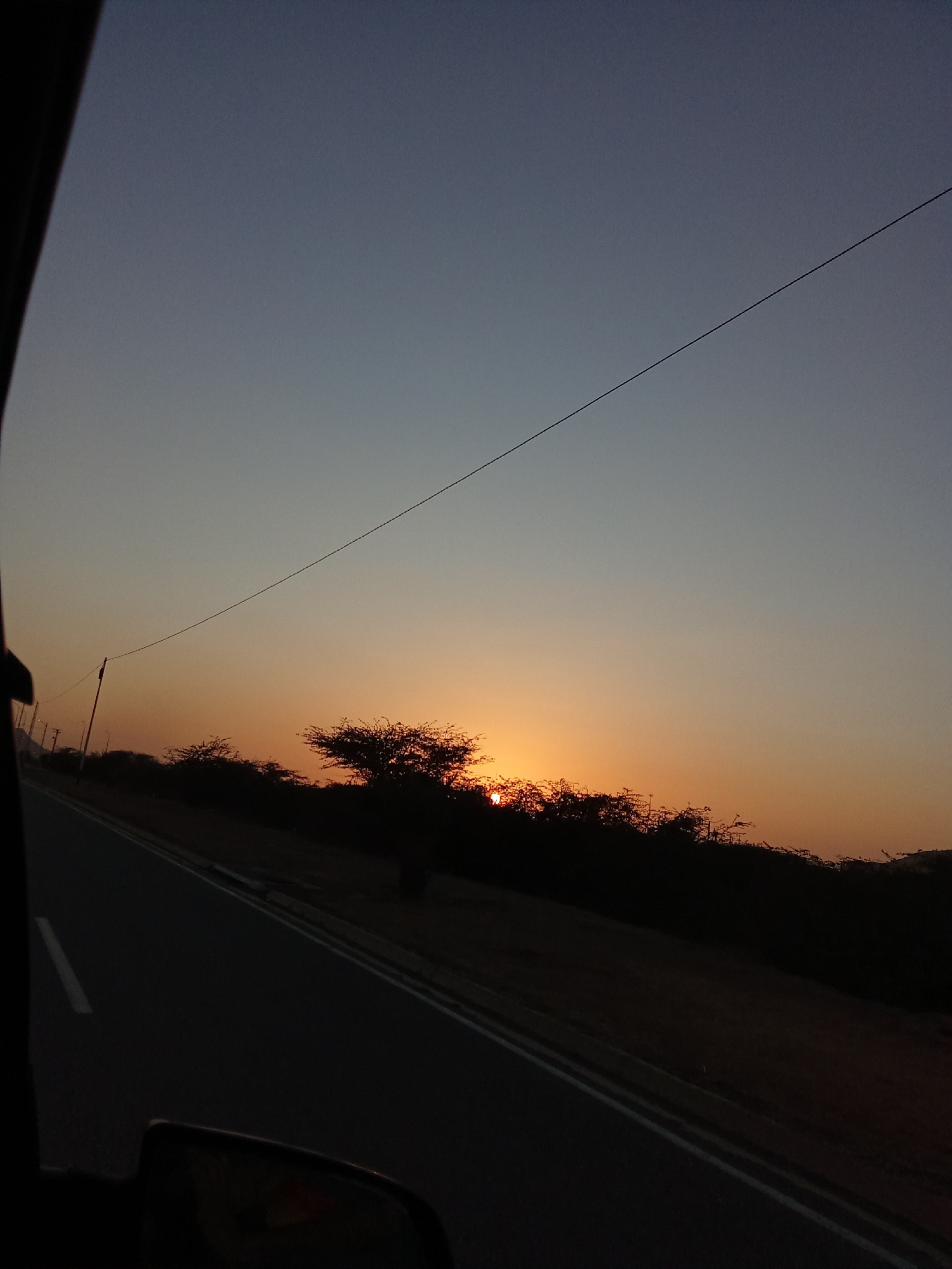 The image depicts a sunset scene, likely taken from inside a moving vehicle. The sky transitions from a pale blue at the top to a vibrant orange-red near the horizon. The sun is setting behind a line of silhouetted trees and shrubs. An asphalt road with a painted white lane marker runs along the left side of the frame. A thin wire or cable stretches diagonally across the upper part of the sky. Parts of the vehicle's interior, including a door frame and side mirror, are visible in the lower left corner, suggesting the photo was taken from inside the car. Juan Griego, Venezuela is not evident from visual elements in the image.