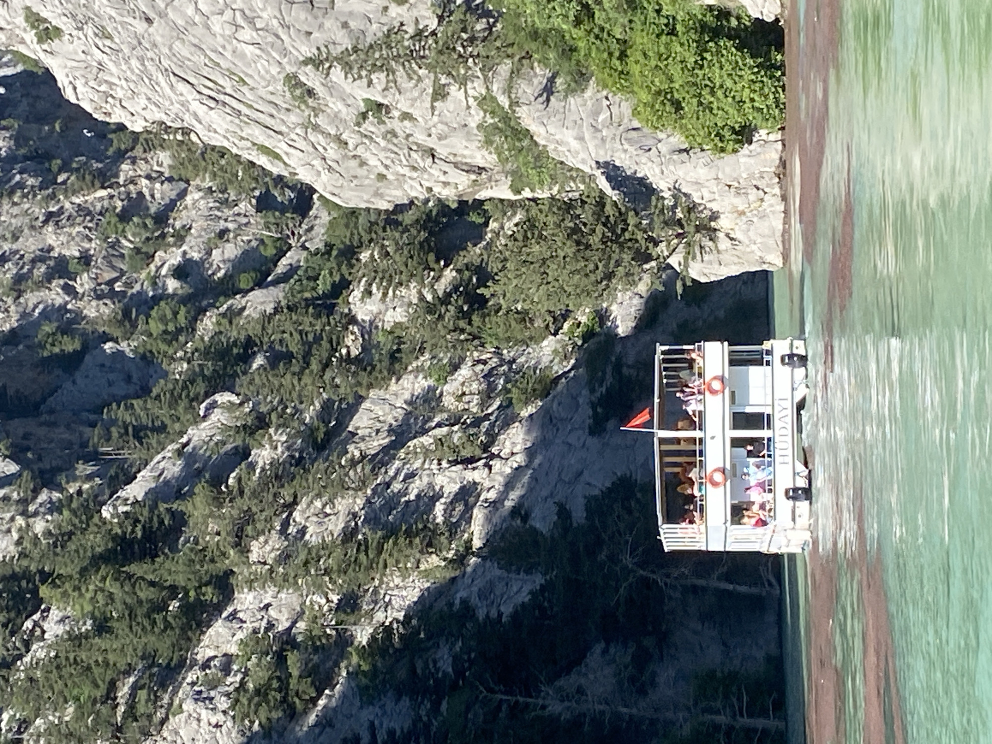 The image, correctly oriented, displays a two-deck tour boat navigating a body of turquoise-green water, likely a lake or reservoir, framed by towering, rocky mountains. The scene is set near Akseki, Turkey.
The mountains, steep and rugged, are composed of light-colored rock and are partially covered with green trees and scattered vegetation. Bright sunlight illuminates the scene, casting shadows that suggest it is daytime, possibly morning or early afternoon. The weather appears clear and sunny.
A white passenger boat, named "HÜDAYİ" on its side, is central to the foreground. Numerous people are visible on both its upper and lower decks, enjoying the cruise. They appear casually dressed, engaged in a leisurely activity, likely sightseeing. A small red flag, possibly the Turkish flag, flies from the boat's upper deck. Two orange lifebuoys are affixed to the railings on the lower deck.
The water exhibits a vibrant turquoise color, interspersed with reddish-brown streaks or reflections, which might indicate mineral content or sediment in the water. The overall impression is one of a tranquil and scenic natural environment, popular for recreational excursions.
