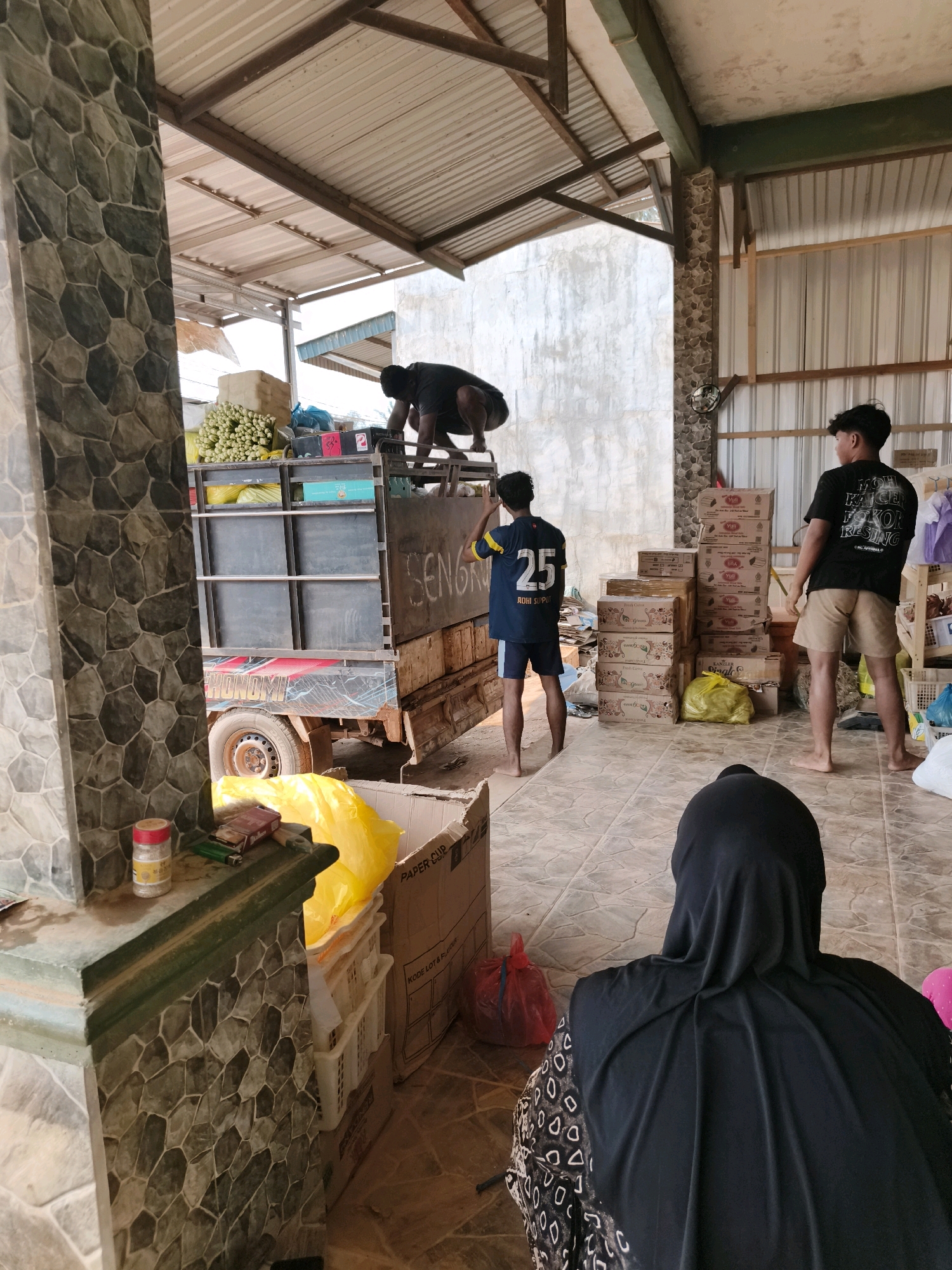 In an outdoor, open-air structure with a corrugated metal roof supported by wooden beams, two young men and another individual wearing a black hijab are present. One man, in a black t-shirt and shorts, is standing near the back of a flatbed truck and looking away from the camera. Another man, wearing a blue and yellow jersey with the number "25" and shorts, faces the truck, his back to the camera. A third individual, obscured by the framing and wearing a black hijab and patterned clothing, is seated, facing away from the camera.
The truck is loaded with goods, including what appear to be bunches of green produce, stacked boxes, and bags. The truck bed has some writing on its side, including "SENGR" which is partially visible. In the foreground, there is a low wall or counter with items on it, including cigarettes and a container of white granules. Cardboard boxes and plastic bags are scattered around the tiled floor. The background features a concrete wall. The overall scene suggests a loading or unloading operation in a commercial or storage area.