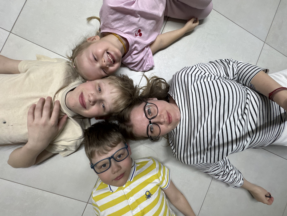 Four individuals are lying on their backs on a light-colored tiled floor, arranged radially with their heads meeting at a central point and bodies extending outwards, in an indoor location within Gornyi (Dvurechenskoe S/P), Russia. An adult female, positioned at the top of the frame, has long brown hair, wears black-rimmed glasses, a black and white striped long-sleeve shirt, and white pants; a small tattoo is visible on her left forearm. To her left is a young girl with blonde hair, wearing a pink t-shirt with a red cherry graphic and a light-colored beaded necklace. To her right is a boy with brown hair, wearing blue-rimmed glasses and a yellow and white striped short-sleeve shirt with a blue logo on the left chest. Below the adult female, at the bottom of the frame, is another boy with blonde hair, wearing a light beige t-shirt, with his right hand resting on his chest. All four subjects are looking directly at the camera with slight to broad smiles.