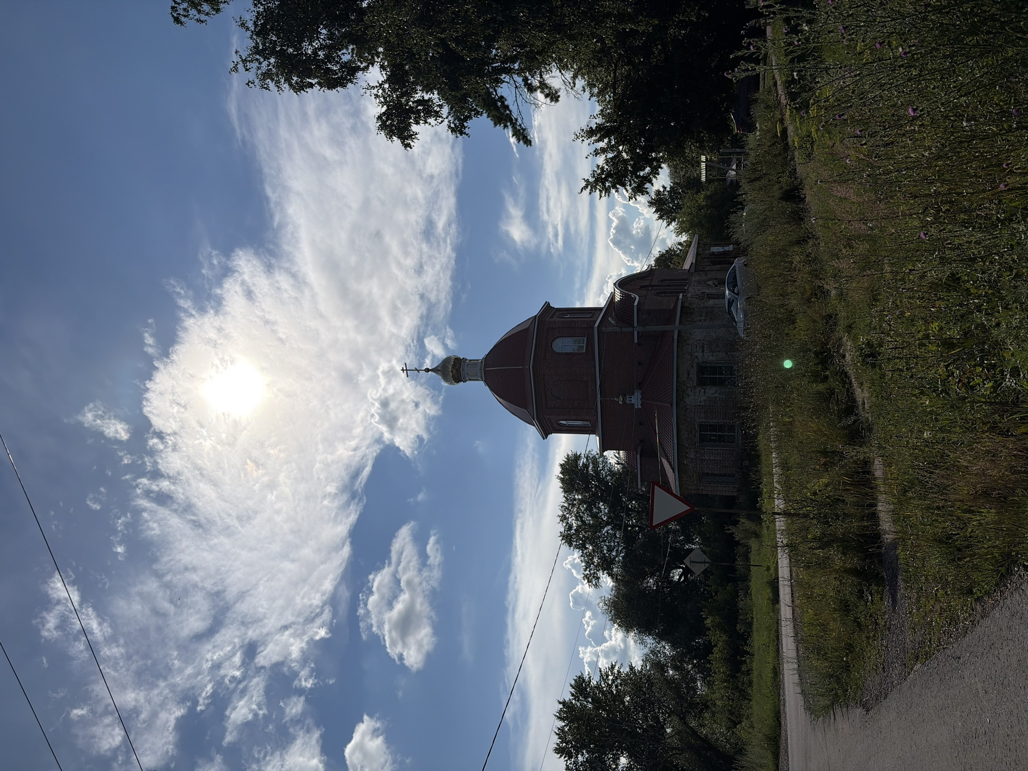 The image shows a brick church with a red dome and a golden cross on top, located in the village of Ryndino, Russia. The sun is shining brightly, partially obscured by scattered clouds. The church is surrounded by trees and vegetation, with a road curving in the foreground. There are also power lines visible in the sky. The scene appears to be peaceful and rural.
