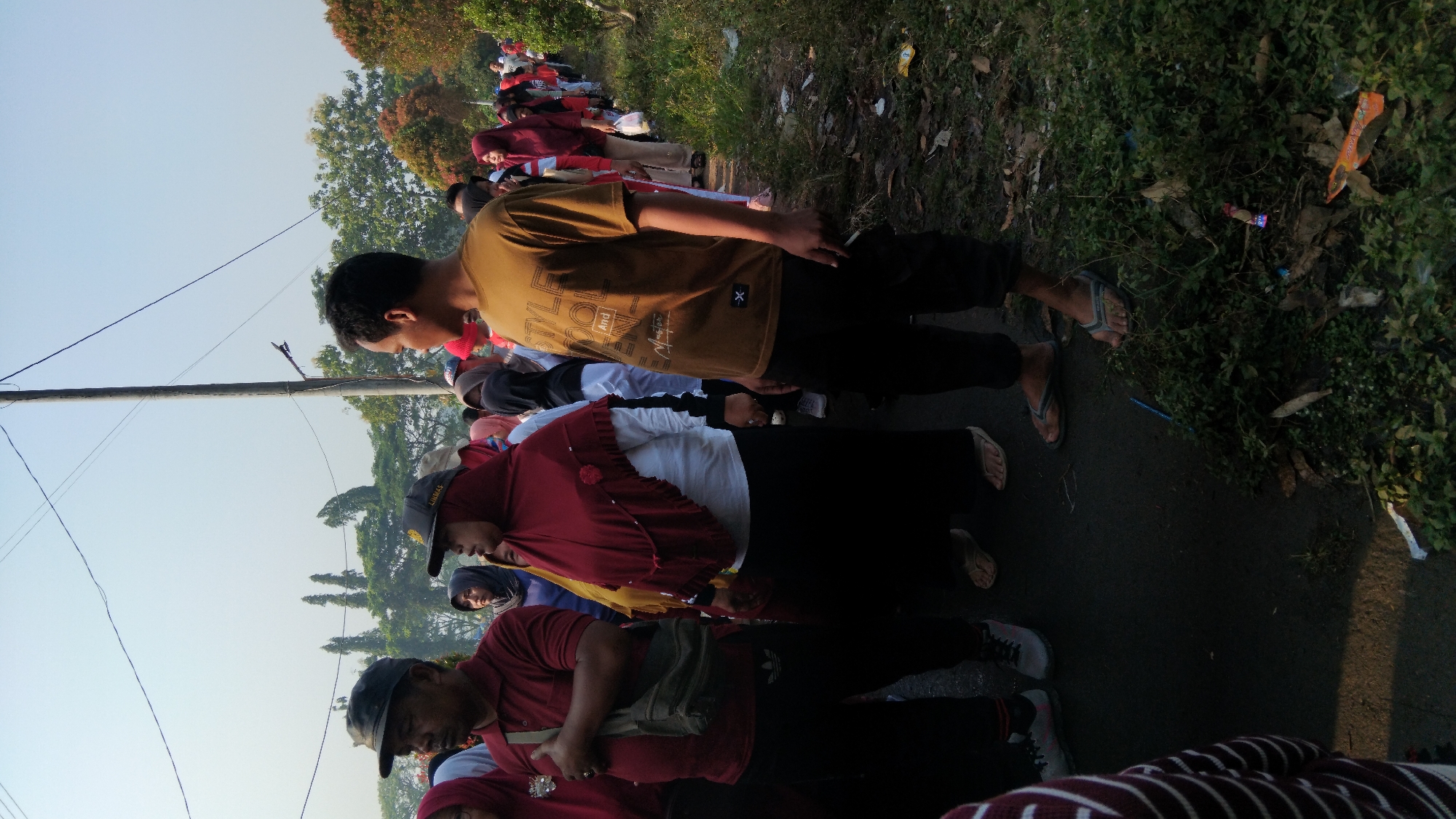 In a daytime outdoor scene in Posong, Indonesia, a diverse group of people are standing in a line, facing towards the right of the frame. The majority of individuals are wearing headscarves, indicative of Muslim attire. The ground is asphalt, with a grassy, overgrown verge to the right featuring scattered litter. A utility pole with electrical wires stretches across the sky, which is a clear, bright blue with a slight haze.

In the foreground, a man wearing a dark cap and a maroon polo shirt stands on the left, with a green canvas bag slung over his shoulder. Beside him, a woman in a maroon headscarf and white top is visible. To the right, a man in a light brown t-shirt with text and black pants stands prominently, looking towards the side. Further into the background, the line of people continues, with various headscarf colors and clothing styles. The trees in the background are lush and green, with some showing hints of autumn colors. The overall atmosphere suggests a public gathering or event.