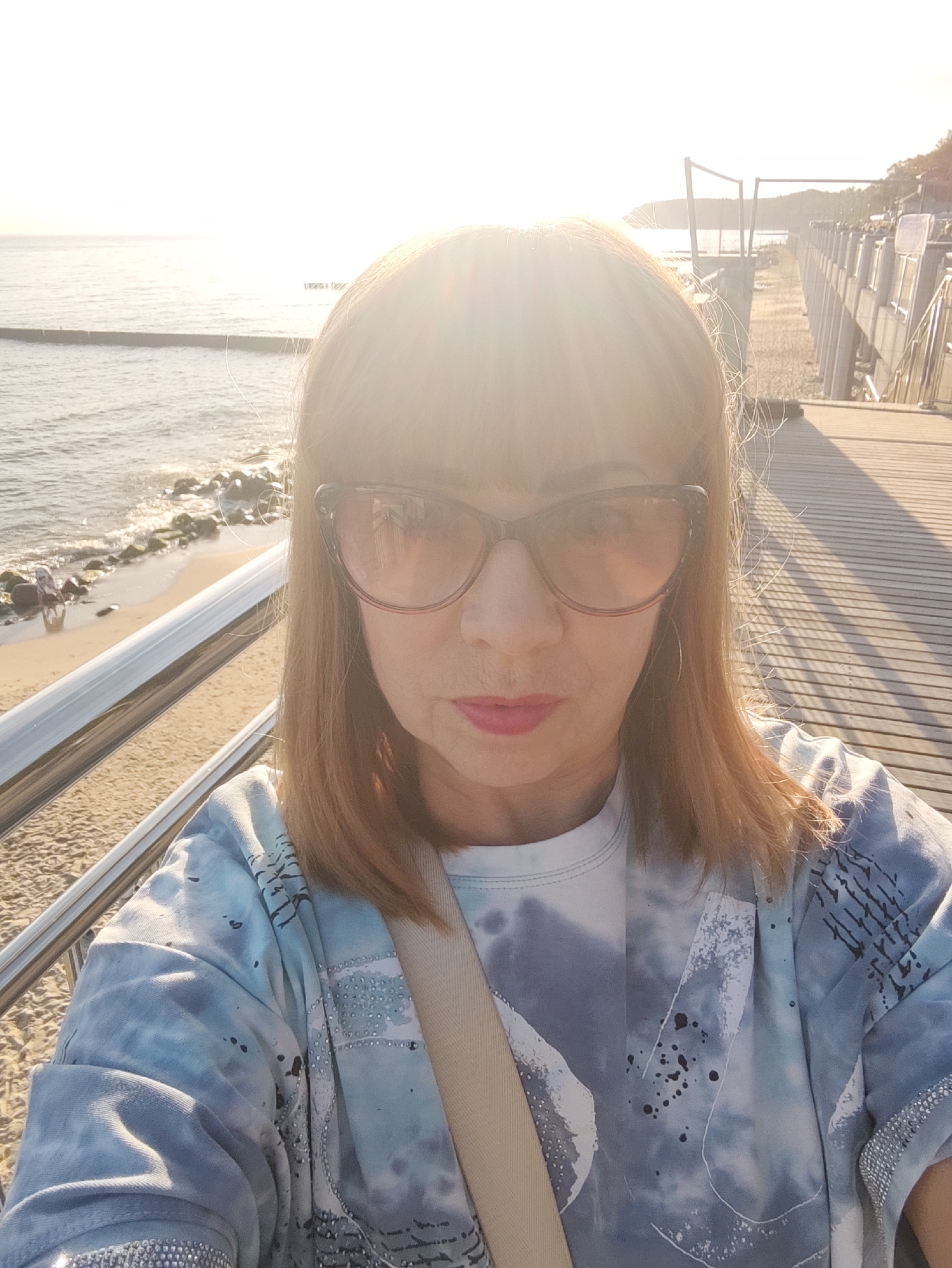 A woman takes a selfie outdoors during the day. She is positioned in the foreground, facing the camera directly, with her head and upper torso visible. She has shoulder-length blonde hair and is wearing large, dark-rimmed sunglasses and a light blue tie-dye t-shirt adorned with silver embellishments along the sleeves. A beige strap is visible across her chest. Her lips are painted with a pink lipstick.
Behind her, a wooden boardwalk stretches into the distance, casting long shadows on its surface. To her left, a beach with sand and rocks lines the edge of the Baltic Sea. The sea is calm, with gentle waves. A breakwater structure is visible in the water. In the far background, a coastline with trees and buildings can be seen, suggesting an urban or resort area. The sun is low in the sky, creating a bright glare that obscures details in the upper portion of the image and illuminates the scene with a warm, golden light. The overall setting appears to be the promenade or a pier in Svetlogorsk, Russia, known for its coastal location on the Baltic Sea.