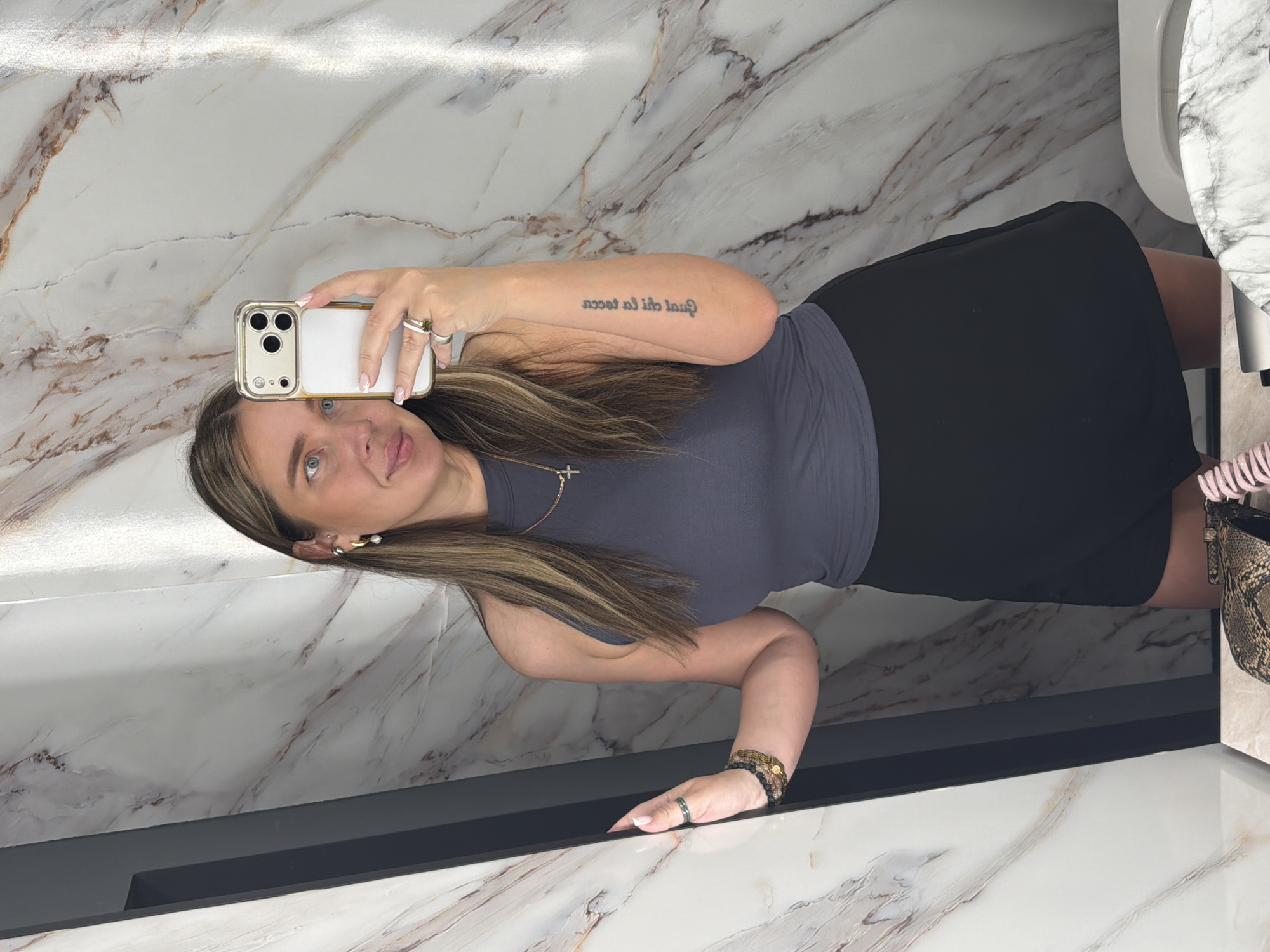 A woman is taking a selfie in a bathroom. She is holding an iPhone in her right hand. She is wearing a grey top and a black skirt. There is a marble countertop and wall behind her. A sink is to her right. The location is unknown.