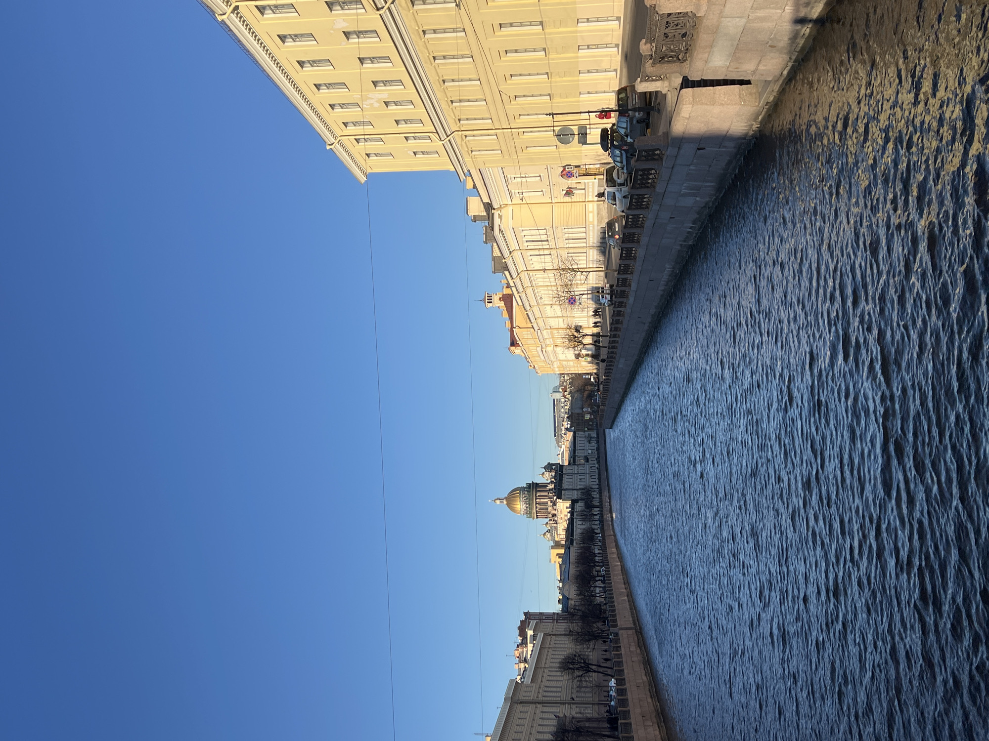 The image depicts a canal in Saint Petersburg, Russia, with buildings lining either side. The canal water is choppy. On the right side, buildings stand adjacent to the water's edge, with parked cars visible along the street. A golden-domed cathedral, likely Saint Isaac's Cathedral, is visible in the distance on the right. Above, the sky is clear and blue.