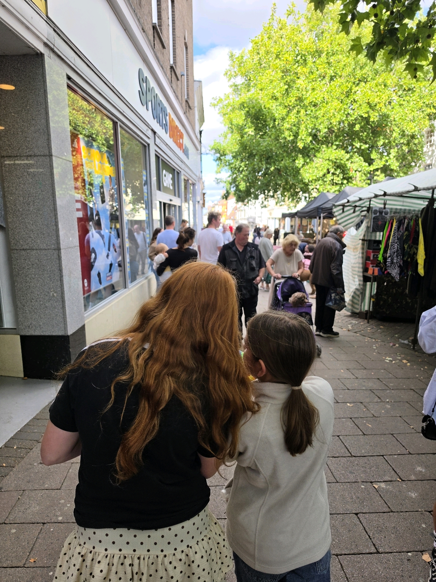 A low-angle, medium shot captures a street scene in West Suffolk, United Kingdom, on a cloudy day. In the foreground, two females, a woman with long auburn hair and a younger girl with brown hair tied in a ponytail, stand facing away from the camera. The woman wears a black t-shirt and a cream-colored polka-dot skirt. The girl is dressed in a light gray fleece jacket and blue jeans. They are standing on a paved walkway, with a building on the left and market stalls on the right.
The building on the left features a large storefront with a gray granite facade and large windows displaying clothing and advertisements. The text "SPORTS DIRECT" is visible on the facade.
To the right, several market stalls are set up along the walkway. One stall, covered by a green and white striped awning, displays clothing on hangers. Another stall is partially visible behind it.
In the background, various people are walking along the street. A man in a dark jacket is seen pushing a stroller with a small dog in it. Other individuals, including a woman in a light-colored top and a man in a black shirt, are also present. Trees with lush green foliage are visible further down the street. The overall impression is a bustling local market or shopping street.
