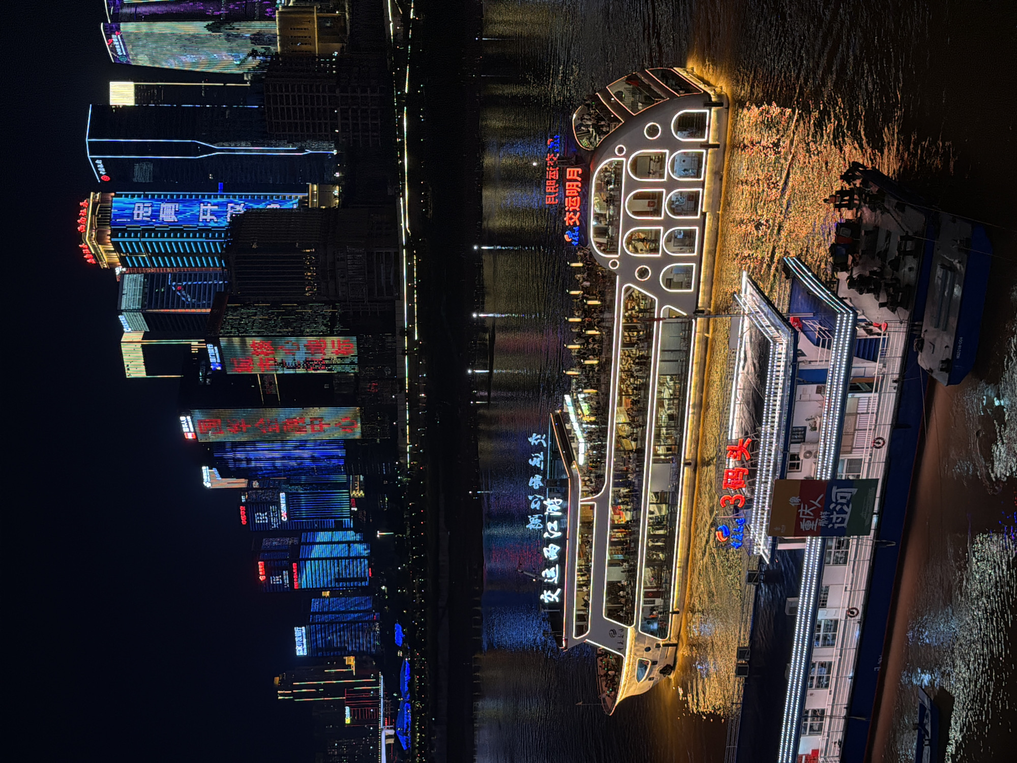 The image depicts a nighttime cityscape in Yuzhong District, China, characterized by a vibrant display of illuminated skyscrapers and a riverfront illuminated by the reflections of city lights and a fleet of illuminated boats. The dominant subjects are two large passenger boats docked in the foreground and mid-ground, respectively, on a body of water that appears to be a river.
The larger, more prominent boat, in the mid-ground, is a multi-decked vessel with a white hull and numerous arched windows, outlined in bright white lights. The upper deck is crowded with people visible through the windows and along the railing, suggesting it is actively carrying passengers. The boat features prominent signage in Chinese characters, illuminated in white. The water surrounding this boat reflects the warm, golden lights of the boat and the distant city.
The boat in the foreground, a lower, more industrial-looking vessel, has a dark blue hull and a white upper structure. It is also lined with lights. A banner with Chinese characters is displayed on its side. Several people are visible on the deck of this boat, some appearing to be workers with cargo. A smaller, dark-colored boat is partially visible to the lower left, floating in the water.
In the background, a dense collection of modern skyscrapers rises, their facades aglow with a dazzling array of colorful lights. The lights on the buildings are in various patterns and colors, including blue, red, and white, creating a dynamic and futuristic urban landscape. The city lights create a shimmering reflection on the dark water, adding depth and visual interest to the scene. The overall atmosphere is one of a bustling and technologically advanced urban environment at night.