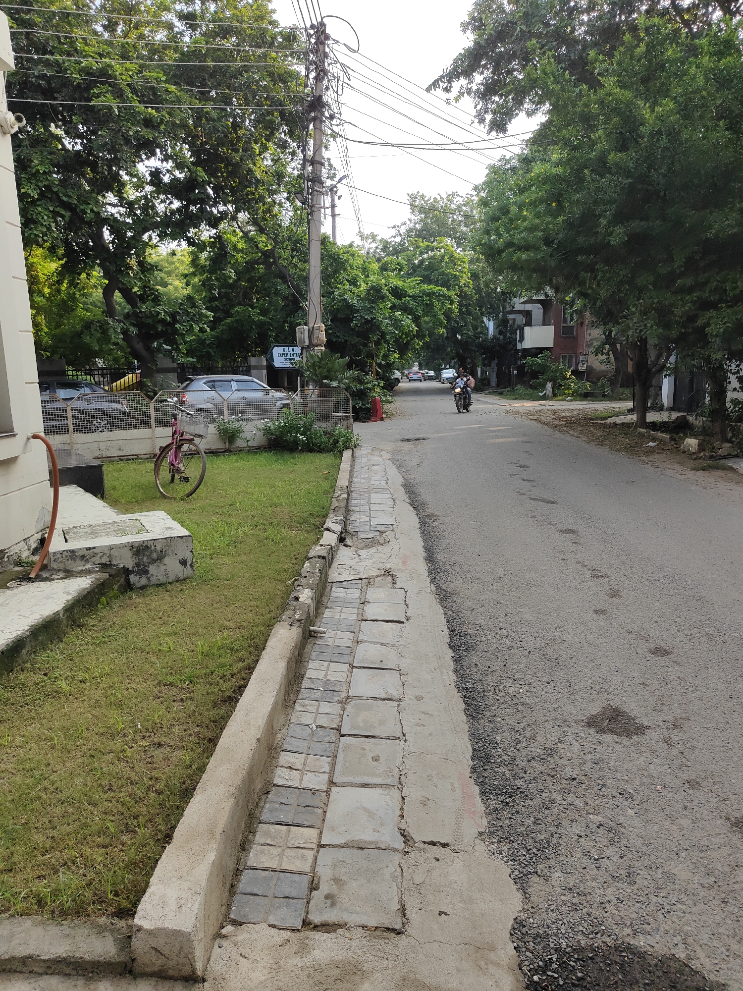 The image depicts a street scene in Gurgaon, India, likely in a residential or mixed-use area. A paved road runs from the foreground into the distance, bordered on the left by a narrow concrete walkway with a tiled surface. Adjacent to the walkway is a patch of grass, on which a pink bicycle is parked. Further left, a portion of a building is visible, along with an orange hose attached to a white pipe. The right side of the road is lined with trees, buildings, and utility poles with numerous wires. In the distance, several cars are parked, and a person is riding a motorcycle on the road. The sky is overcast.