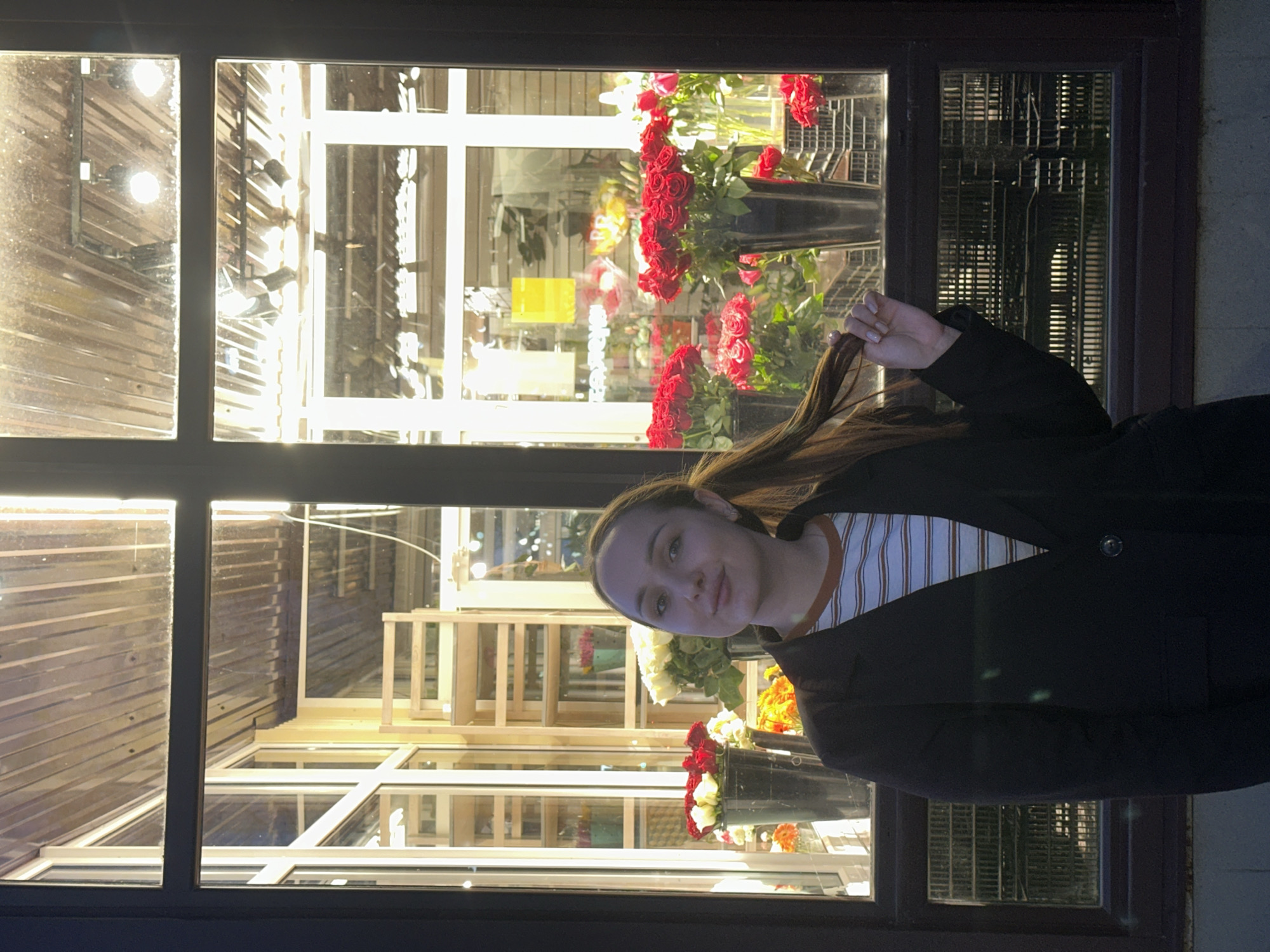A young woman stands in front of a window display of a flower shop. The window is divided into multiple panes. Inside the flower shop, red and white flowers are arranged in containers. There are also wooden shelves visible. The location is Chaikovsky, Russia. The young woman is wearing a striped shirt and a dark coat. She is holding a strand of her hair.
