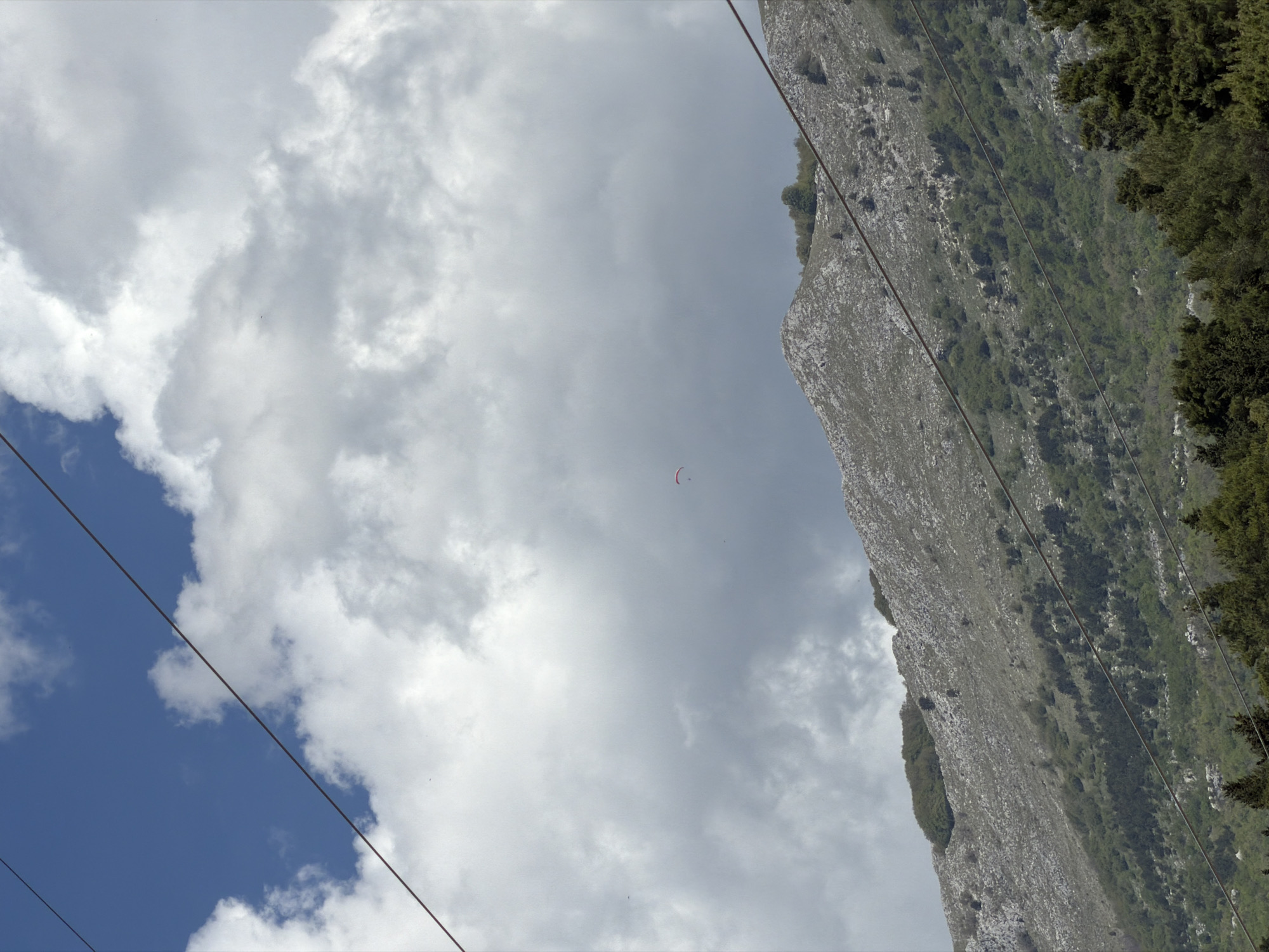 This image, when correctly oriented, showcases a scenic mountainous landscape under a bright, partly cloudy sky, characteristic of Budva, Montenegro. The upper portion of the frame is dominated by a dynamic interplay of expansive white cumulus clouds and patches of clear blue sky.
A central subject of the photograph is a single, small red paraglider canopy, visibly soaring through the sky, indicating an individual engaged in paragliding. Below the sky, rugged mountainsides rise, characterized by grey rock formations interspersed with dense patches of green trees and shrubbery.
Two prominent, thin dark lines, likely wires (possibly power lines or part of a cable car system), stretch diagonally across the scene from the lower left to the upper right. The bright daylight and clear visibility suggest fair weather conditions. There is no visible text in the image. The scene captures an active outdoor pursuit within a dramatic natural setting.