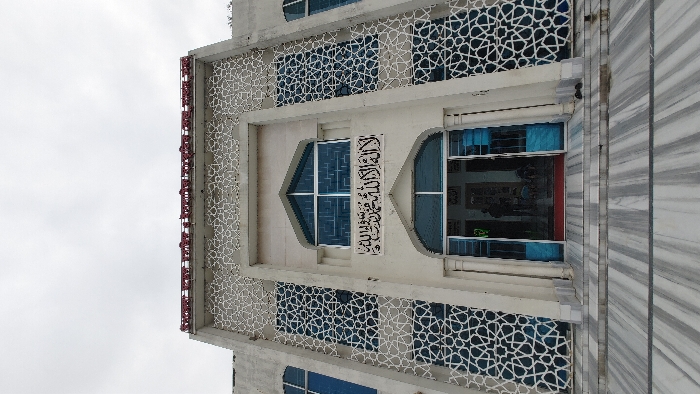 This is a low-angle photograph of the facade of a multi-story building, likely a mosque, associated with Bangladesh Open University, as indicated by a prominent red Bengali inscription reading 'বাংলাদেশ উন্মুক্ত বিশ্ববিদ্যালয়' (Bangladesh Open University) on its upper facade. The structure is situated in the Savar area of Bangladesh.
The building features a light-colored (off-white/light grey) exterior with symmetrical design. Its central section includes an arched main entrance with open, blue-framed glass double-doors. Above the entrance, a rectangular black plaque displays Arabic calligraphy, reading "La ilaha illallah, Muhammadun rasulullah" (There is no god but Allah, Muhammad is His messenger). Directly above this plaque is another arched window with blue-tinted glass.
The facade is extensively decorated with intricate white, abstract geometric patterns that form a screen over blue-tinted windows and wall sections. The building is accessed via wide, horizontally striped grey and white marble steps leading to the entrance. A pair of black shoes are placed on the marble steps to the right of the open doorway. Inside the open entrance, indistinct dark silhouettes of multiple individuals are visible. The sky overhead is uniformly overcast and light grey.