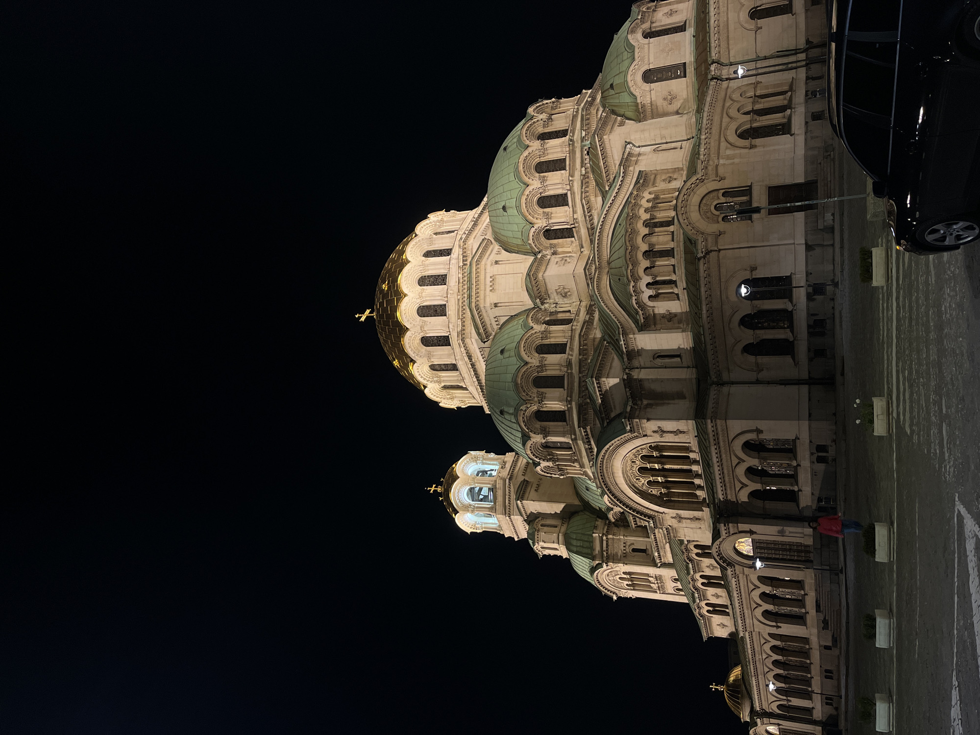 This night photograph, taken in Sofia, Bulgaria, prominently features the Alexander Nevsky Cathedral illuminated against a dark, starless sky. The grand Neo-Byzantine structure dominates the scene, showcasing its distinctive architecture. The largest central dome gleams in gold, topped with an Orthodox cross, while numerous smaller domes are covered in green tiles, adding a striking contrast. The cathedral's elaborate facade is brightly lit, highlighting intricate details such as arched windows, decorative columns, and ornate cornices. A bell tower, also illuminated, is visible towards the left side of the main dome.
At the ground level, paving stones and several white rectangular planters are visible. On the right side of the frame, a dark-colored car, possibly a sedan or SUV, is partially parked, with its front bumper and wheel visible. Towards the lower right, a solitary figure wearing a red jacket stands near the base of the cathedral, offering a sense of the building's immense scale. The clear night provides a stark backdrop, enhancing the dramatic illumination of the cathedral. The scene is quiet, emphasizing the architectural majesty of the building. There is no visible text in the image.