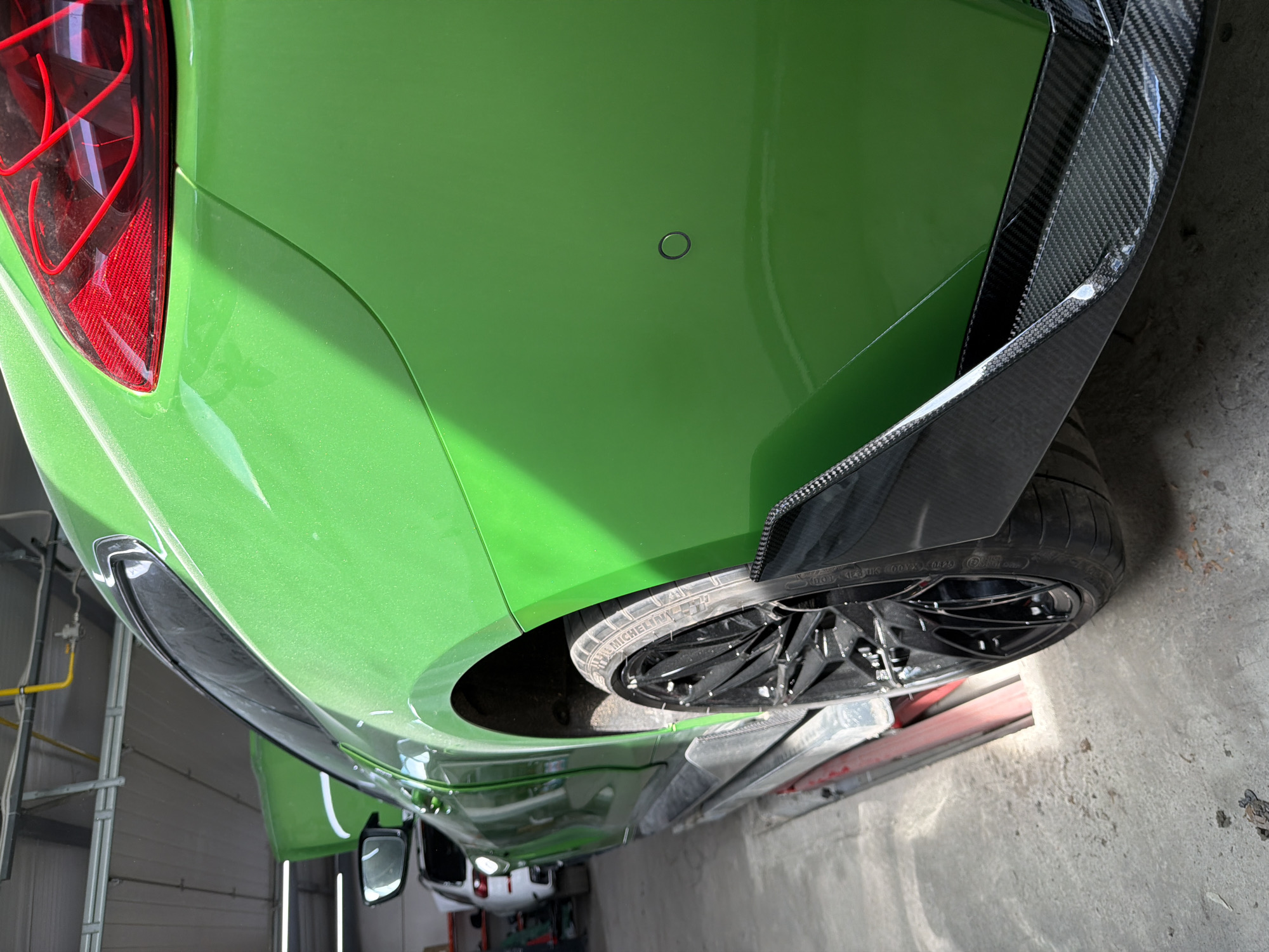 The image shows the rear corner of a bright green car, possibly in a repair shop or garage. The car features a carbon fiber accent and a black wheel with a Michelin tire. In the background, a glimpse of a white car and garage equipment can be seen. The location within Burgas, Bulgaria is not discernible from the visual information provided.