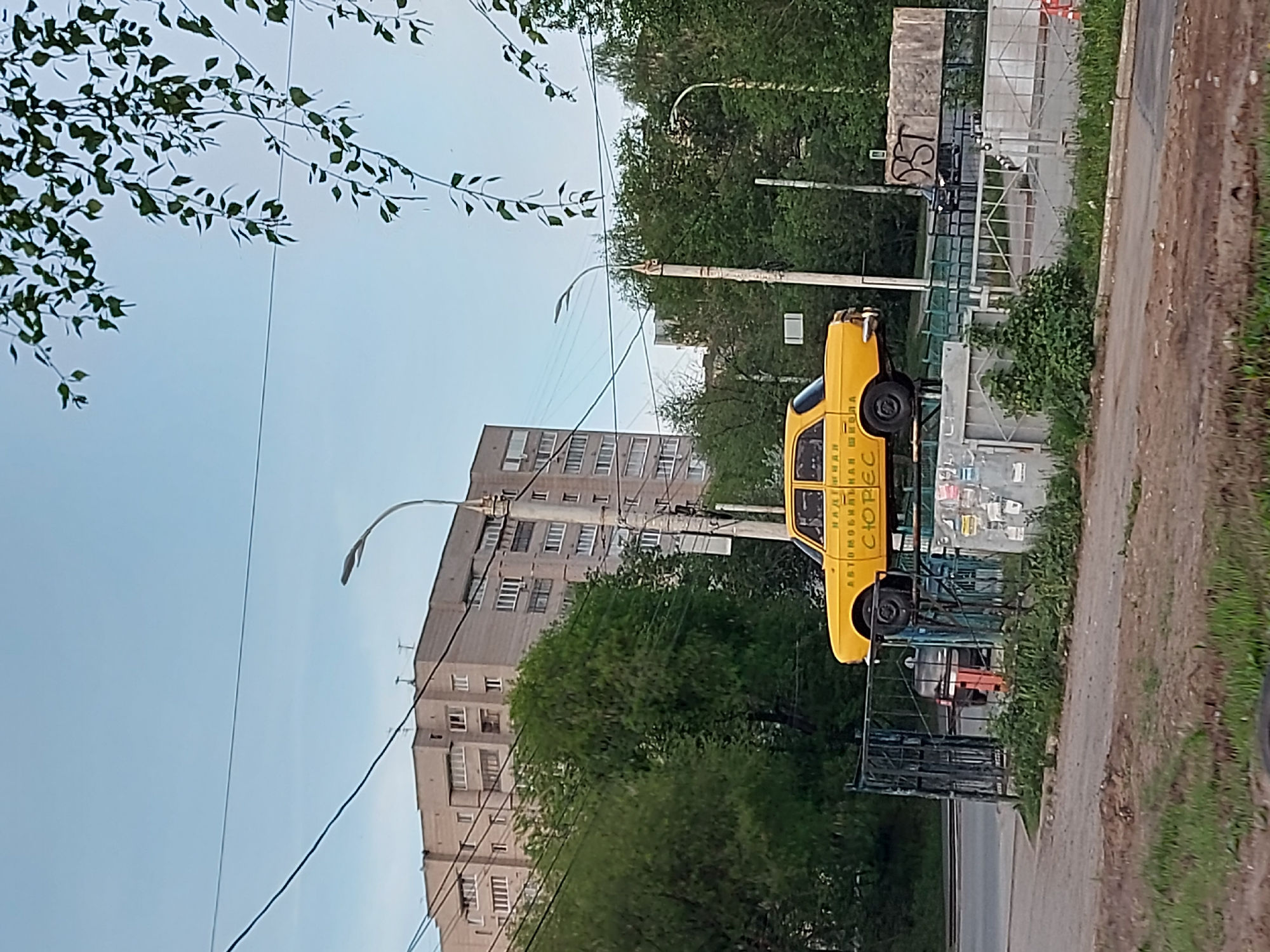 The image shows a yellow car advertisement in Izhevsk, Russia, mounted on a structure beside a road. The car is elevated and displays text on its side. Behind the car, there is a multi-story residential building and numerous trees. Overhead power lines are visible in the foreground. The street and sidewalk are situated to the right of the advertisement. The environment includes a clear sky, indicating daytime.
