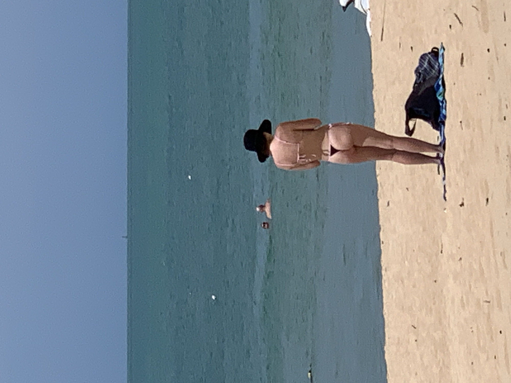 A woman wearing a black wide-brimmed hat and a string bikini stands on a sandy beach, facing away from the camera and towards the ocean. Her bikini is light pink on top and a dark thong on the bottom. Her hands are clasped in front of her. A dark blue and plaid bag lies on a striped beach towel behind her. In the background, two people are swimming in the turquoise water. The sky is clear and blue, with a faint hazy line on the horizon where the sea meets the sky. The scene is likely on a beach in Ko Samui District, Thailand.
