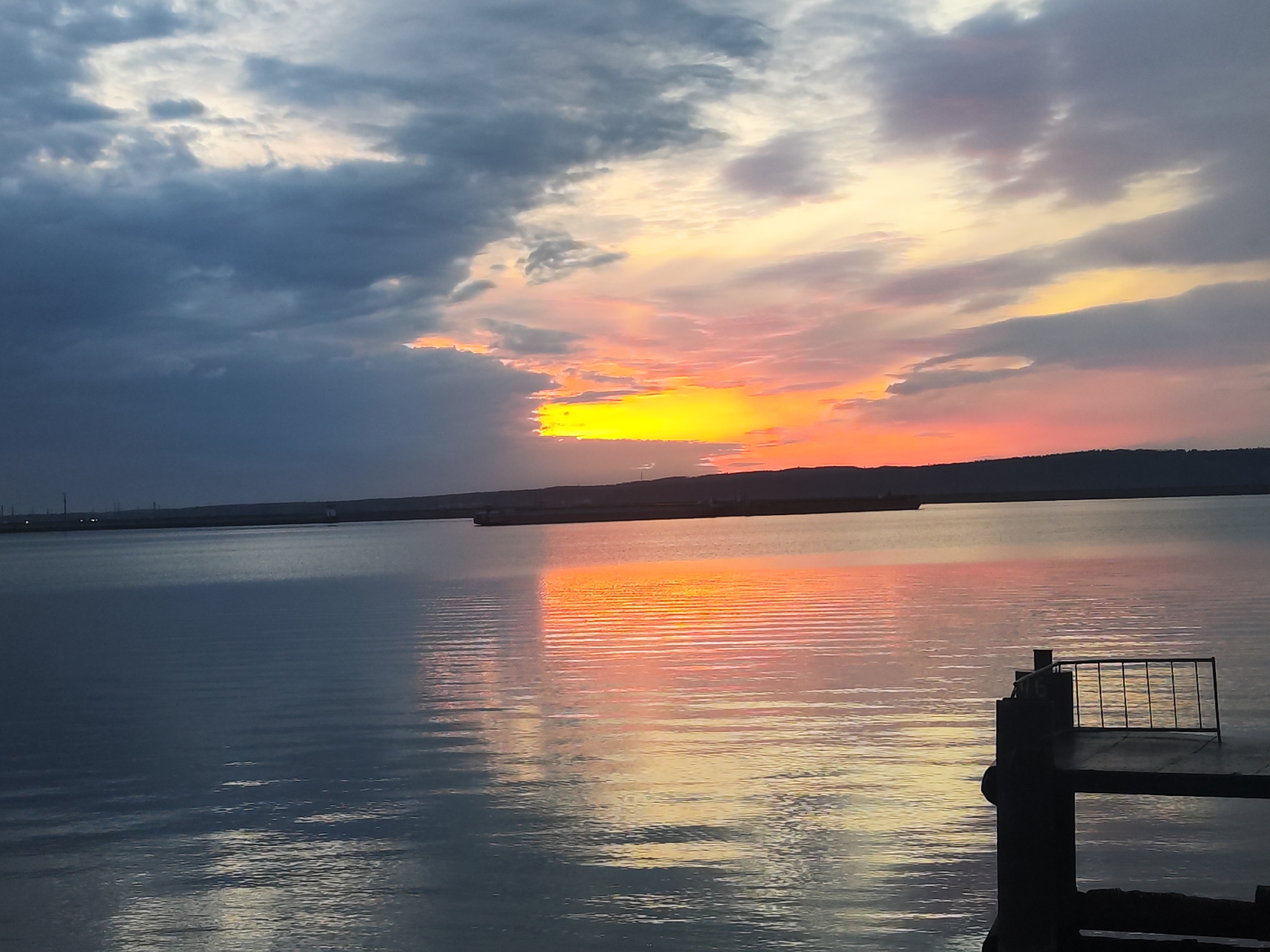The image shows a sunset over a body of water, likely the Kama River, in Chaikovsky, Russia. The sky is a mix of dark, gray clouds and vibrant colors of orange, yellow, and pink where the sun is setting. The water reflects the colors of the sunset, creating a shimmering effect. In the distance, there is a dark shoreline with what appear to be industrial structures and possibly some lights. In the foreground, there is a dark, partially silhouetted structure, possibly a section of a pier or platform, with metal railings. The water has gentle ripples. There are no people visible.