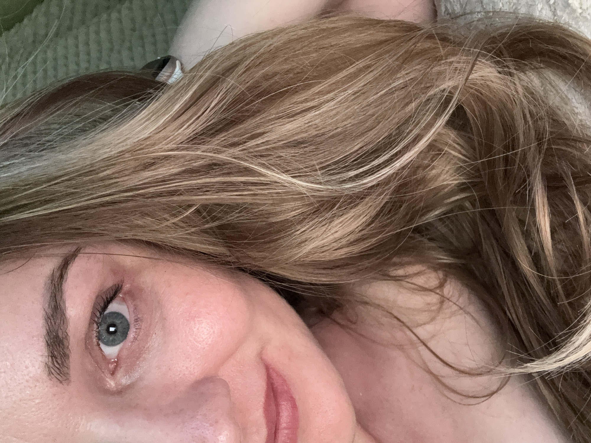 A close-up, shoulder-up shot of a young Caucasian woman with blonde hair, taken from chest level. Her face is angled to the right, with her right eye and the left side of her mouth visible. The background is blurred, with a textured, light-colored surface, possibly a bedspread or blanket. The woman appears to be lying down or reclining. There are no discernible landmarks or elements that would indicate a specific location within Chaikovsky, Russia.