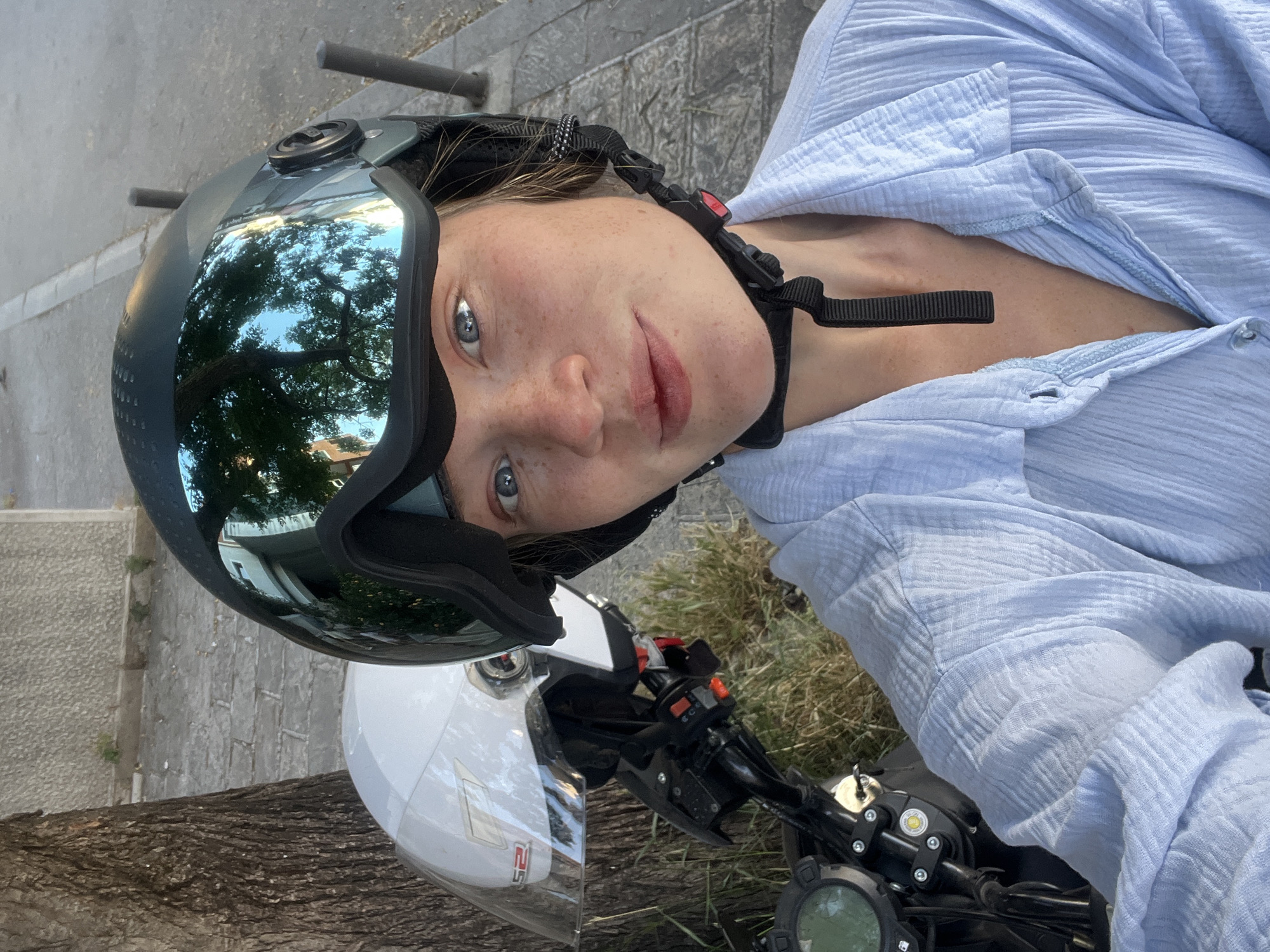 A woman is seated on a motorcycle. She is wearing a blue shirt and a helmet with a reflective visor. The helmet reflects trees and a building. Another white helmet is placed beside the black motorcycle. A dashboard of the motorcycle is visible. The setting is a paved urban area. The location is likely the city of Budva, Montenegro.
