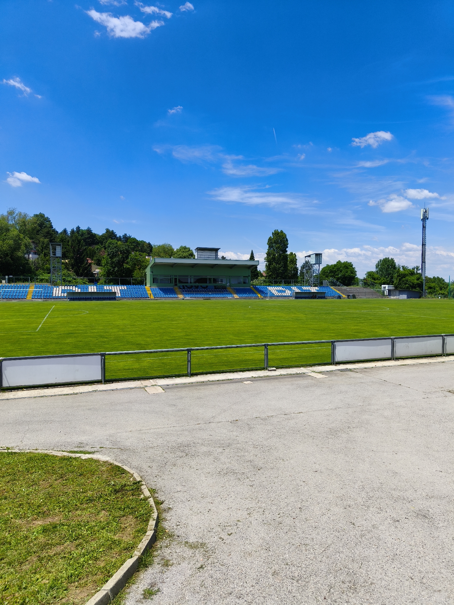The image shows an outdoor football stadium with a well-maintained green grass field. Bleachers with blue and white seating are visible along the sides of the field, with a covered structure behind them. A tall telecommunications tower stands to the right of the stadium, against a bright blue sky with scattered white clouds. Trees and buildings are visible in the background. The scene appears to be set in the city of Sofia, Bulgaria.