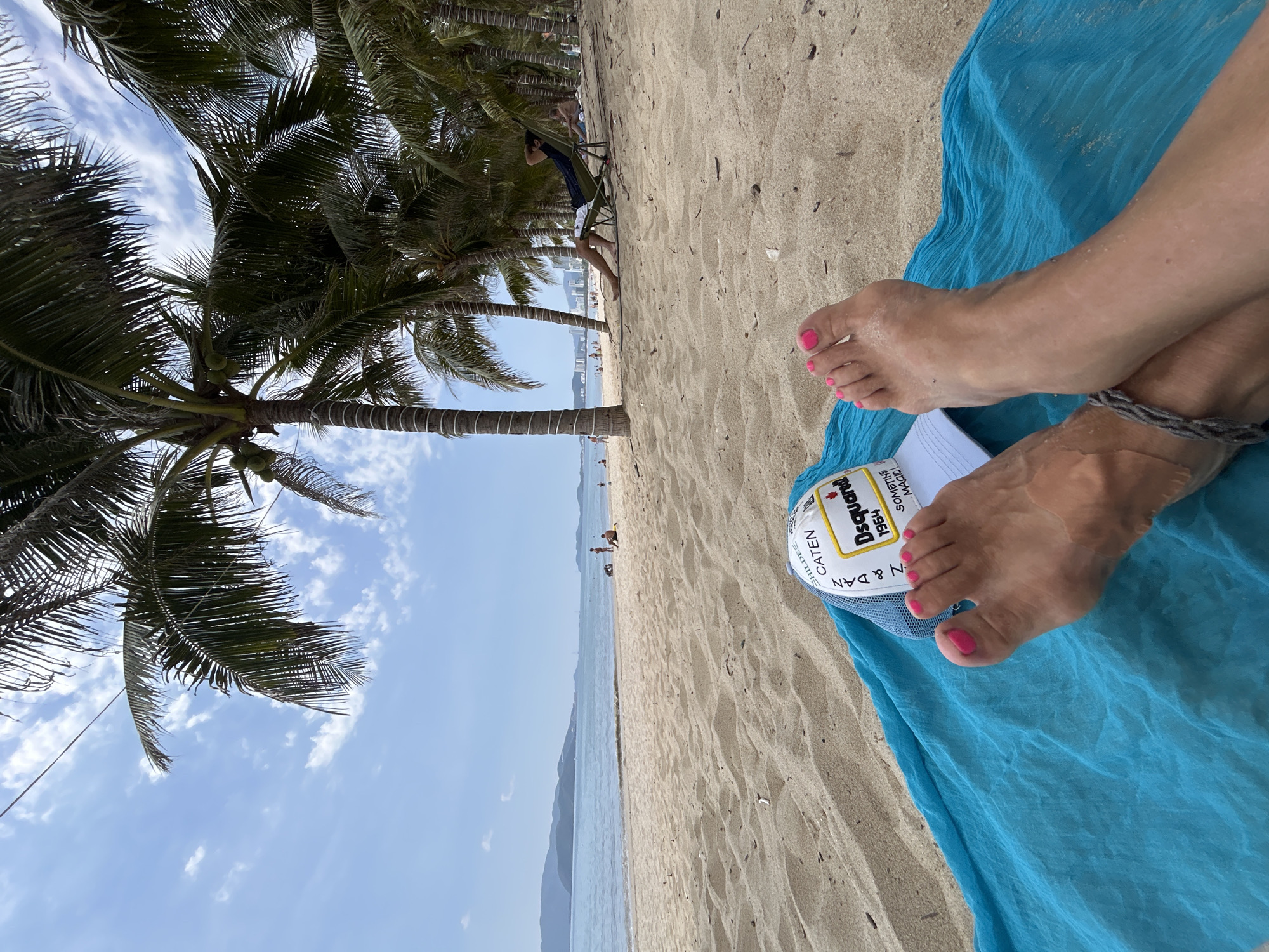 The image captures a beach scene in Nha Trang, Vietnam. A person is lying on a bright blue beach towel, with their bare feet visible, toes painted pink. A white baseball cap with a logo that includes the text "Dsquared" rests between their feet and the towel. The beach is sandy and dotted with palm trees; some people are visible along the shoreline in the distance. The sky is mostly blue with some clouds, and the sea stretches out to the horizon, with a coastal mountain range visible.