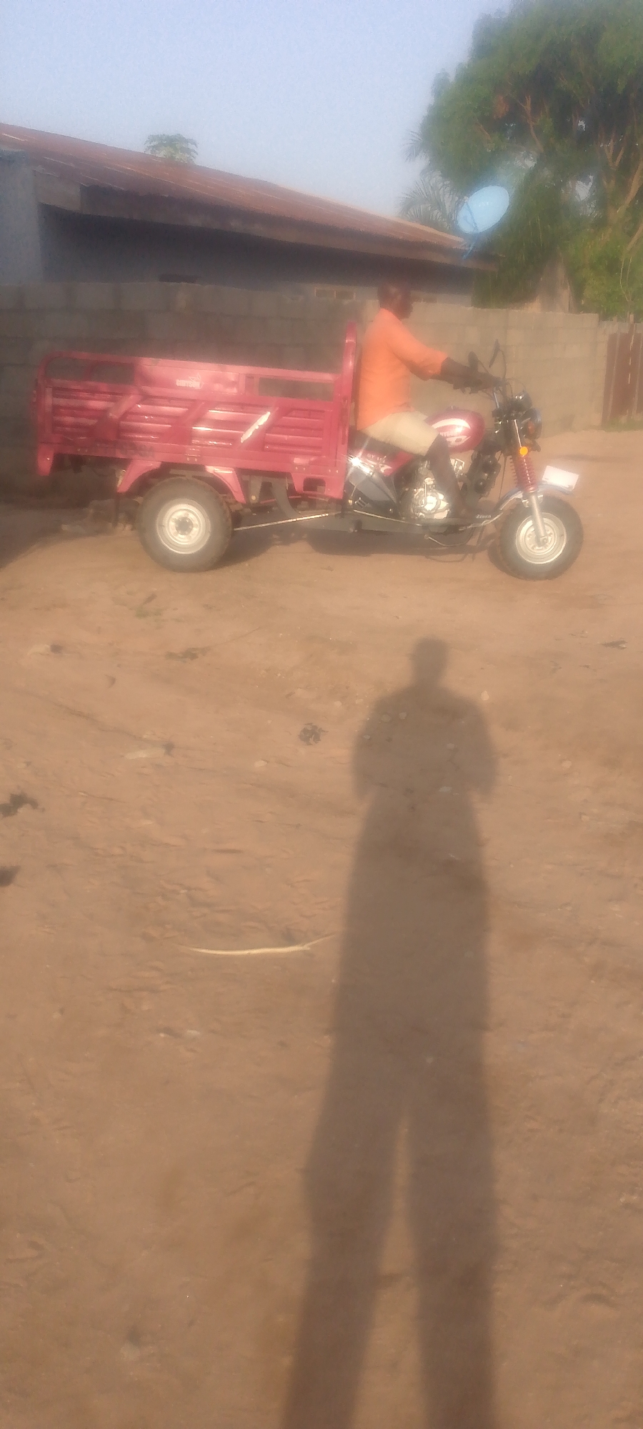 In Jalingo, Nigeria, a man wearing an orange shirt and light-colored pants rides a red three-wheeled cargo motorcycle. The motorcycle has a large cargo bed in the back and a single wheel in the front. The scene appears to be outdoors, with a building with a corrugated metal roof and a brick wall in the background. A satellite dish is visible on top of the building. The ground is covered in dirt and sand, and the shadow of a person is cast on the ground in the foreground. The sky is clear and blue, suggesting it is daytime.