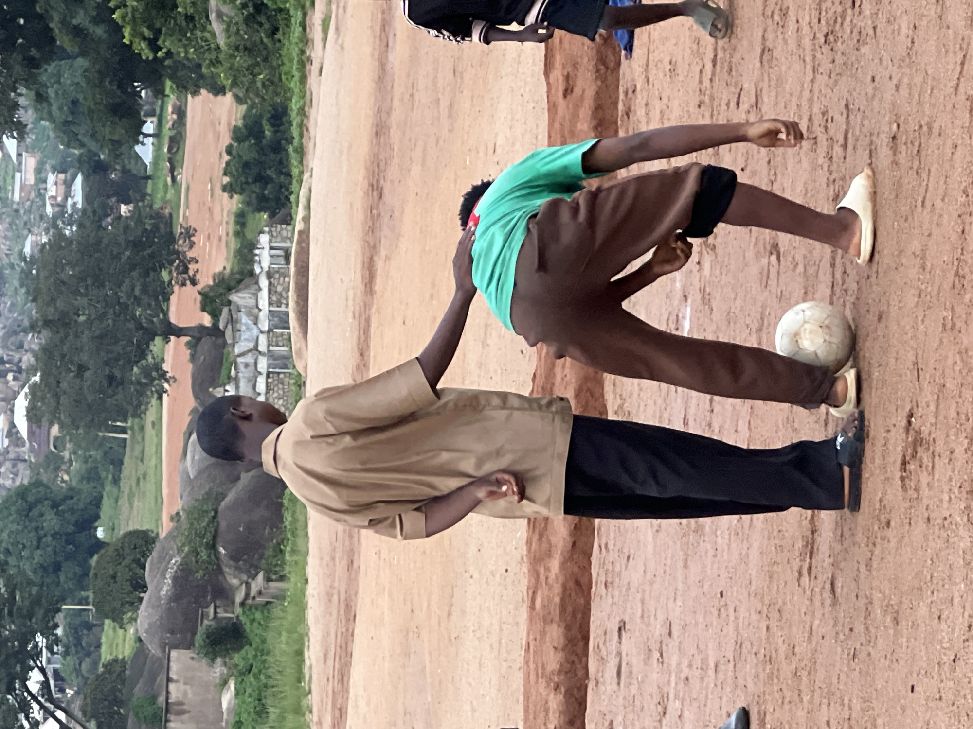 In the city of Jos, Nigeria, two young boys are engaged in a football game on a dusty field. The boy on the left, wearing a tan short-sleeved shirt and black trousers, places a hand on the shoulder of the boy on the right. The boy on the right, in a green t-shirt and brown shorts, is bent over, facing away from the camera, with a white football at his feet. In the background, a hilly landscape with scattered rocks and trees can be seen, with some buildings visible on the slopes. Another boy, partially visible on the right side of the frame, is also present. The environment suggests a rural or semi-urban area with a dirt ground and sparse vegetation.