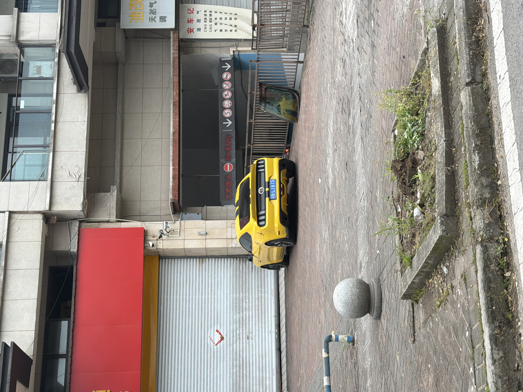 The image shows a yellow sports car parked on a brick-paved street in an urban environment, with a partially open garage door behind it. The car’s license plate, "B-L187S", is visible. To the left of the garage is a red painted area. On the left is a building with glass windows. A street sign displays regulations related to vehicles, including "No Entry" and information about gas and oil conversions. The location is in the city of Chongqing High-tech Zone, China, as indicated by the presence of the Chinese language on the sign. A gray spherical bollard is near the bottom of the frame, and a curb with vegetation runs along the right side of the image. The overall impression is a typical urban scene, possibly a commercial area, with details suggesting a focus on vehicle services.