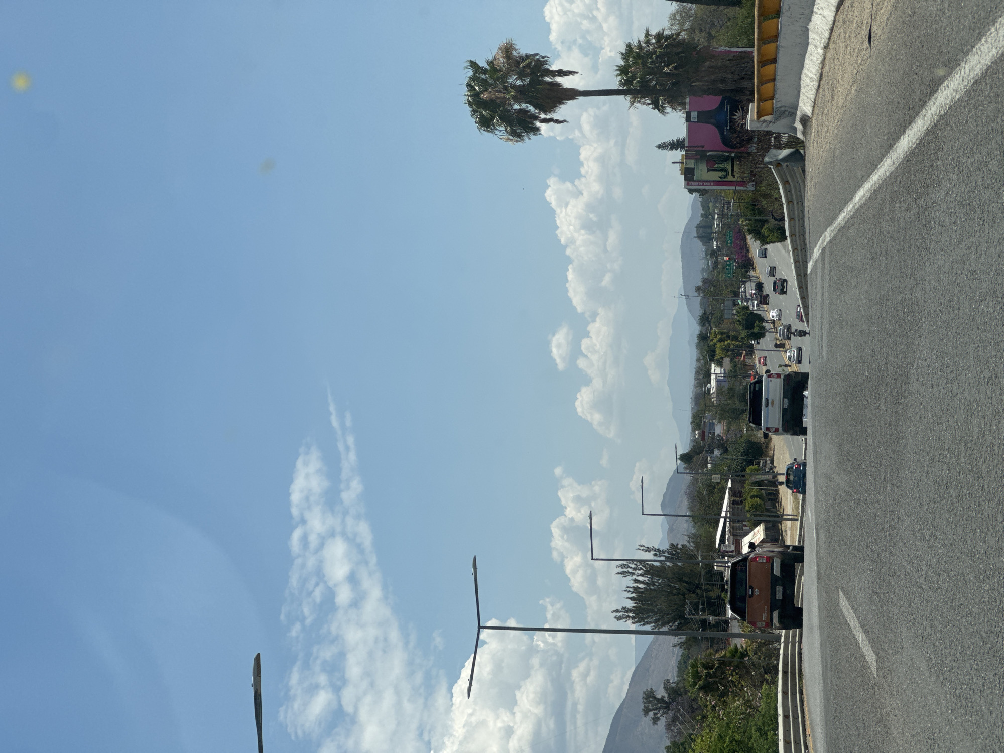 The image depicts a road scene in Tlalixtac de Cabrera, Mexico, under a partially cloudy sky. A two-lane road is visible, with vehicles including cars and a bus. In the background, a cityscape extends towards distant mountains. The foreground features roadside vegetation and streetlights. Palm trees stand along the roadside. A building with pink facade is also visible.