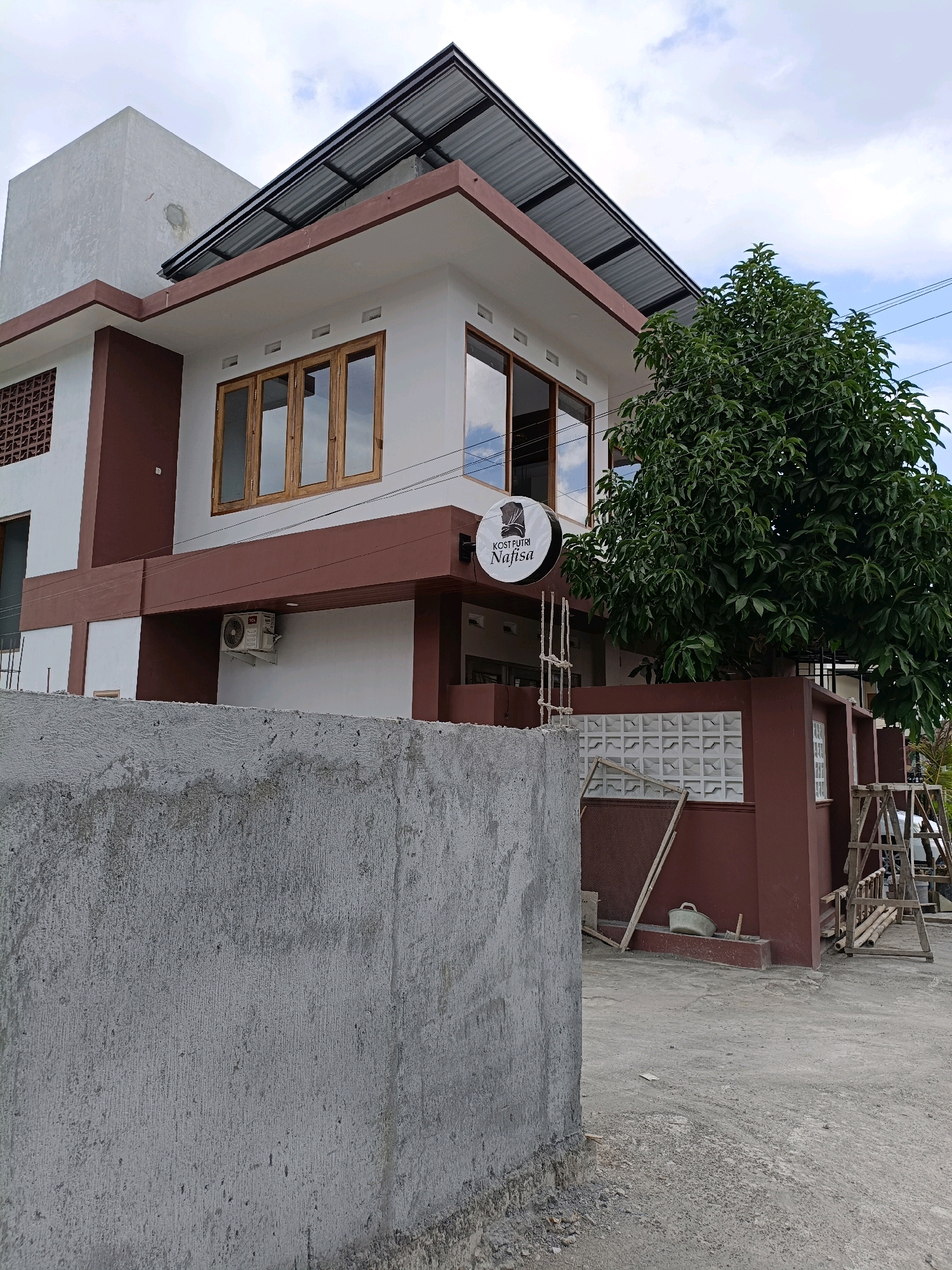 A low-angle, eye-level outdoor photograph captures a two-story building with a modern design in Maguwoharjo, Indonesia. The building is painted in contrasting white and a reddish-brown color, with a multi-pitched corrugated metal roof. Several large, wooden-framed windows are visible, reflecting the sky and clouds. A circular sign with Arabic script and an image of a woman in a hijab is mounted on the exterior wall. An air conditioning unit is attached to the lower section of the building. A large, leafy green tree stands to the right of the building, partially obscuring it. The foreground is dominated by a rough, concrete wall under construction, obscuring the ground level of the building. Construction materials, including wooden planks and scaffolding, are scattered around the property. The sky is partly cloudy with patches of blue. No people are visible in the image.