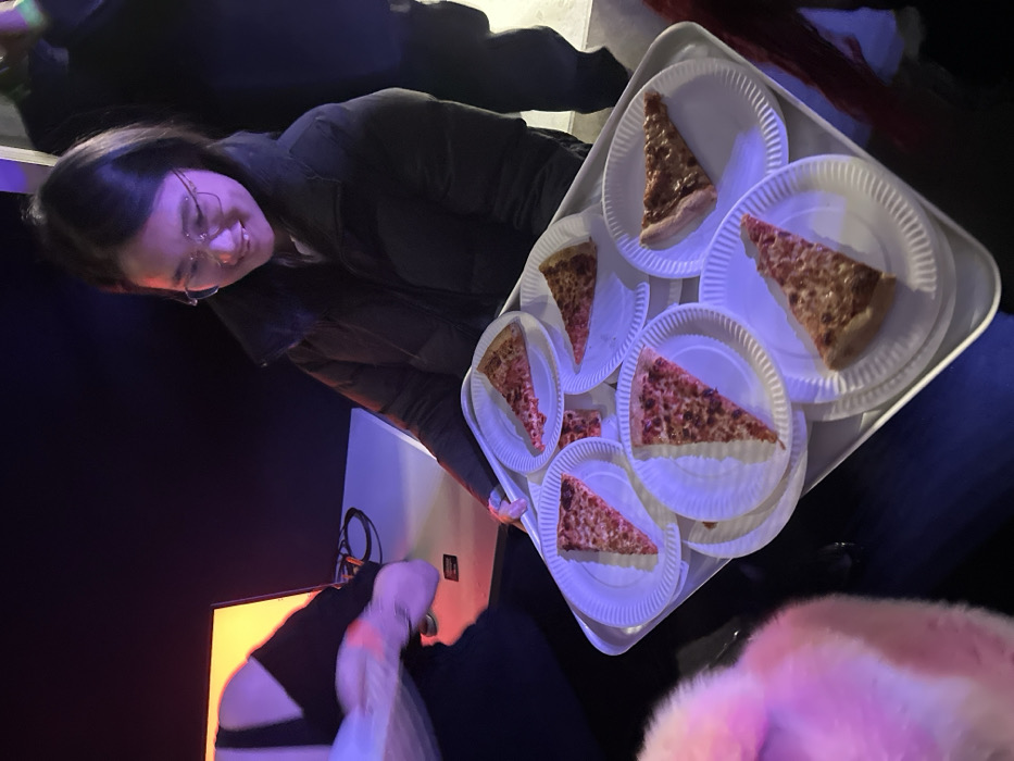 A young East Asian female, estimated to be in her mid-20s, with dark hair and glasses, is positioned centrally in a dimly lit indoor environment. She is smiling and wears a dark puffer jacket. Her hands support a large, white rectangular serving tray holding at least ten individual slices of cheese pizza, each presented on a white, ridged paper plate. A blue-purple light source illuminates the tray and the central figure from the left. To her left, a blurred individual in dark clothing is partially visible, interacting with a white table that features an orange-lit screen and visible cables. Another person, dressed in dark clothing, is partially visible to the right. The general setting suggests an event or social gathering. The precise location within the city of London, United Kingdom, is not discernible from the visual information provided.