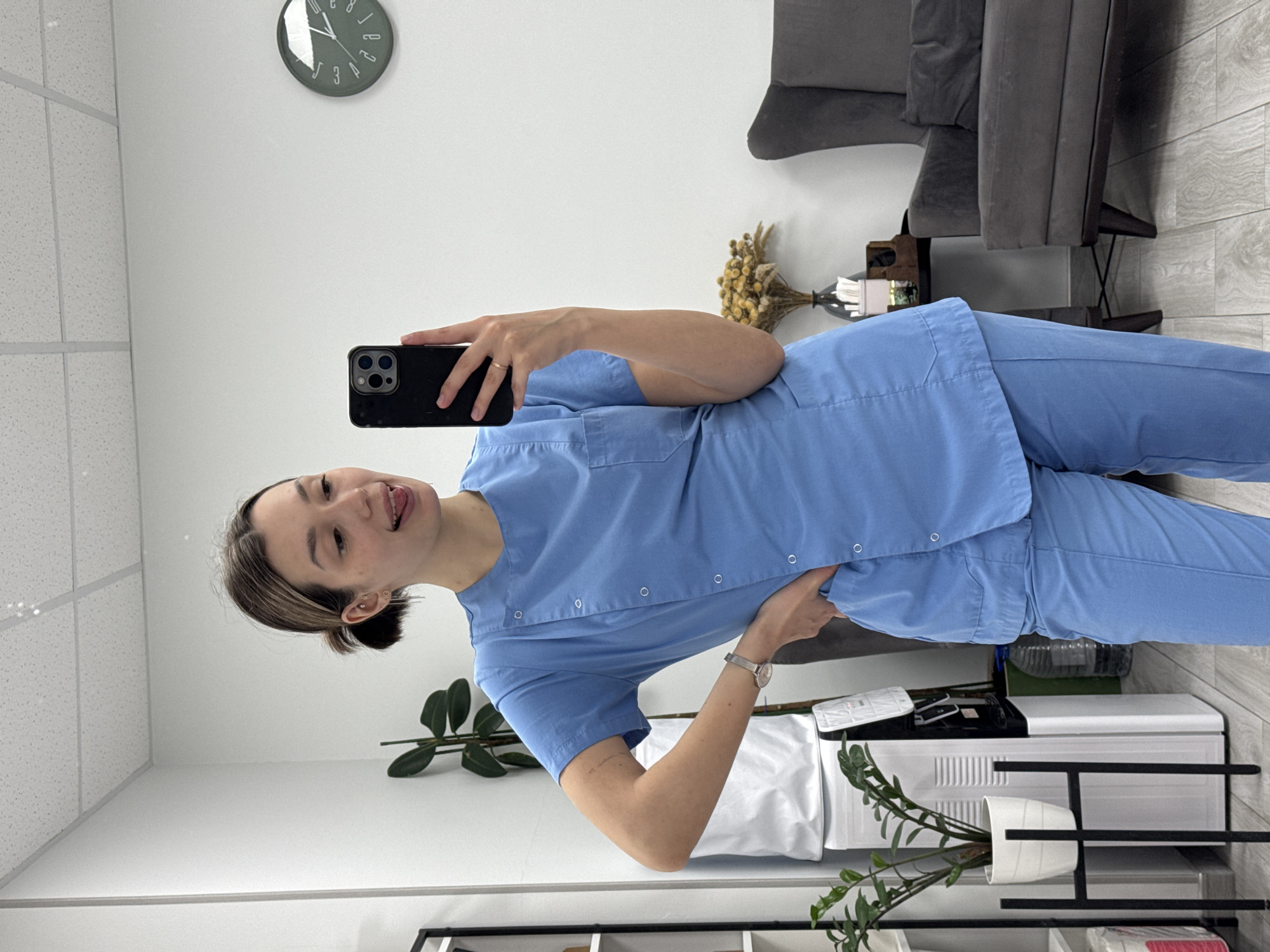 A young adult woman is captured in a mirror selfie, standing upright in what appears to be an indoor office, clinic, or waiting room. She has short, dark brown hair with lighter highlights, pulled back into a bun. She is wearing light blue medical scrubs, comprising a short-sleeved top with white snap buttons and matching pants. Multiple small earrings adorn her left ear, and a silver-toned watch is on her left wrist.
She holds a black smartphone with a prominent triple-lens camera array in her right hand to take the photo, while her left hand rests on her hip. Her expression is playful and cheerful, as she smiles broadly and sticks out her tongue.
The background features plain white walls and a white drop ceiling with grid panels. The floor is light-colored wood or laminate. To her right, a dark gray upholstered armchair or sofa is visible. Behind her, on a small table, there is a vase with a bouquet of dried, light-colored flowers and a white container, possibly a tissue box. A large green leafy plant in a white pot is situated near a white appliance or cabinet, with a black shelving unit partially visible below. A circular green wall clock with white numbers (1-12) is on the upper left wall, showing the time is approximately 2:40 or 2:50. No other discernible text is visible.