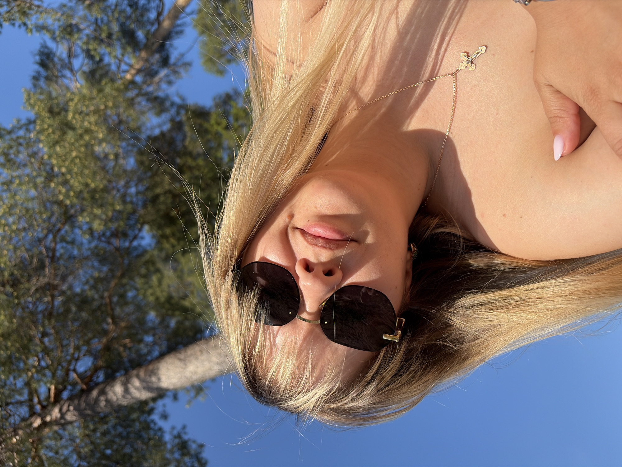 A woman with blonde hair, wearing sunglasses and a gold cross necklace, is positioned against a backdrop of blue sky and a tree with green foliage. The location cannot be confirmed as Chaikovsky, Russia, without additional information.