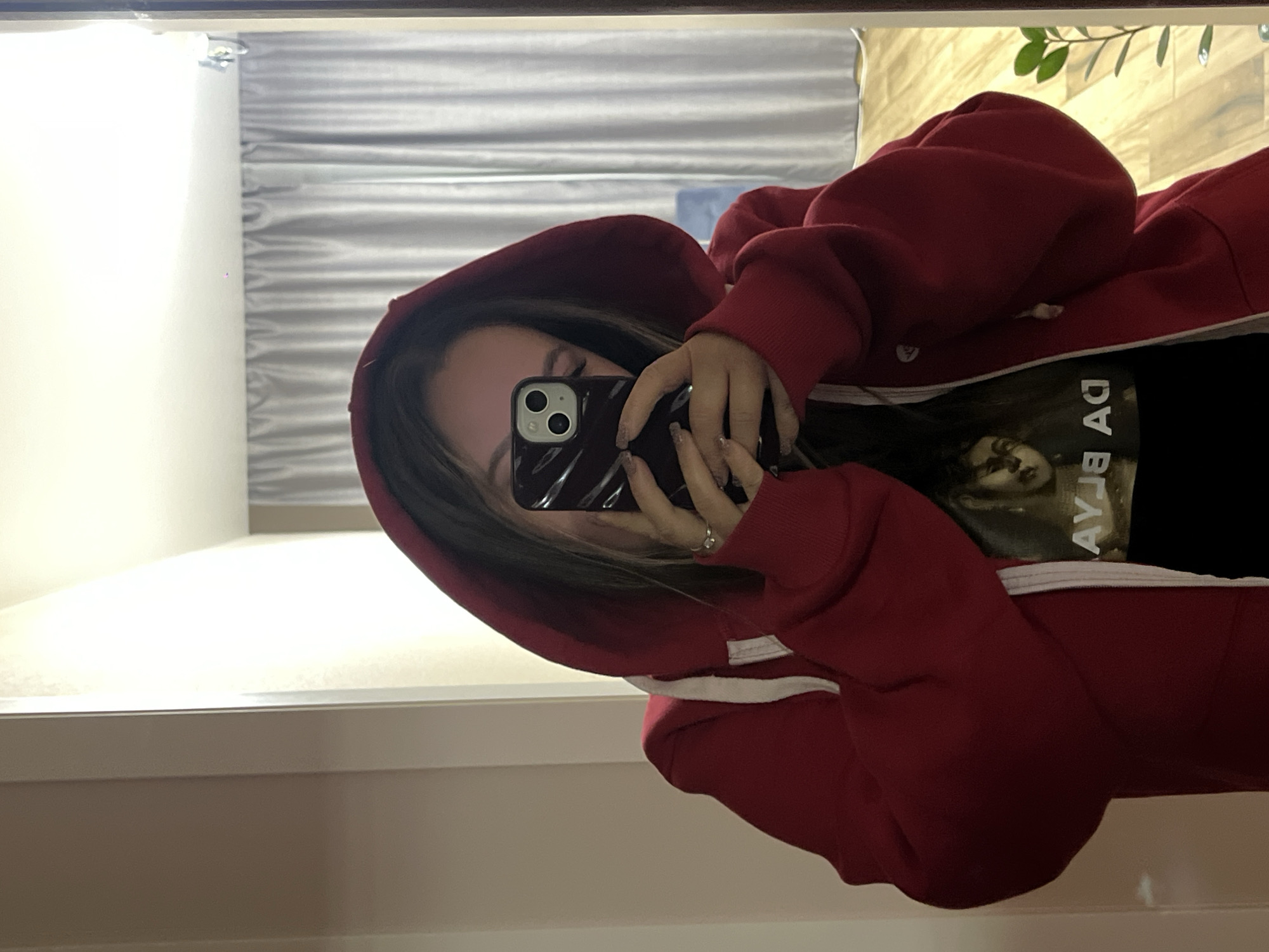 The image depicts a young woman taking a mirror selfie indoors. She is seen from the chest up, wearing a red hooded sweatshirt with the hood up, over a dark t-shirt. She holds a black smartphone, featuring a dual-lens camera system, which largely obscures her face. A ring is visible on her right hand, and her fingernails have polish or a design.
The activity taking place is a mirror selfie, where the subject captures her reflection using her phone.
Notable details include the indoor setting, which appears to be a room. The background, visible in the mirror's reflection, consists of a white wall or vertical structure to the left, and light-colored horizontal blinds or curtains behind the subject. A reflection of green plant leaves can be seen in the upper right corner of the mirror. The lighting suggests an indoor setting, possibly daytime or evening with artificial light. On her dark t-shirt, an inverted graphic design is visible, featuring a face and partially discernible letters that appear as "AY J8" when read as they are reflected (from right to left, upside down). The overall scene is well-lit.