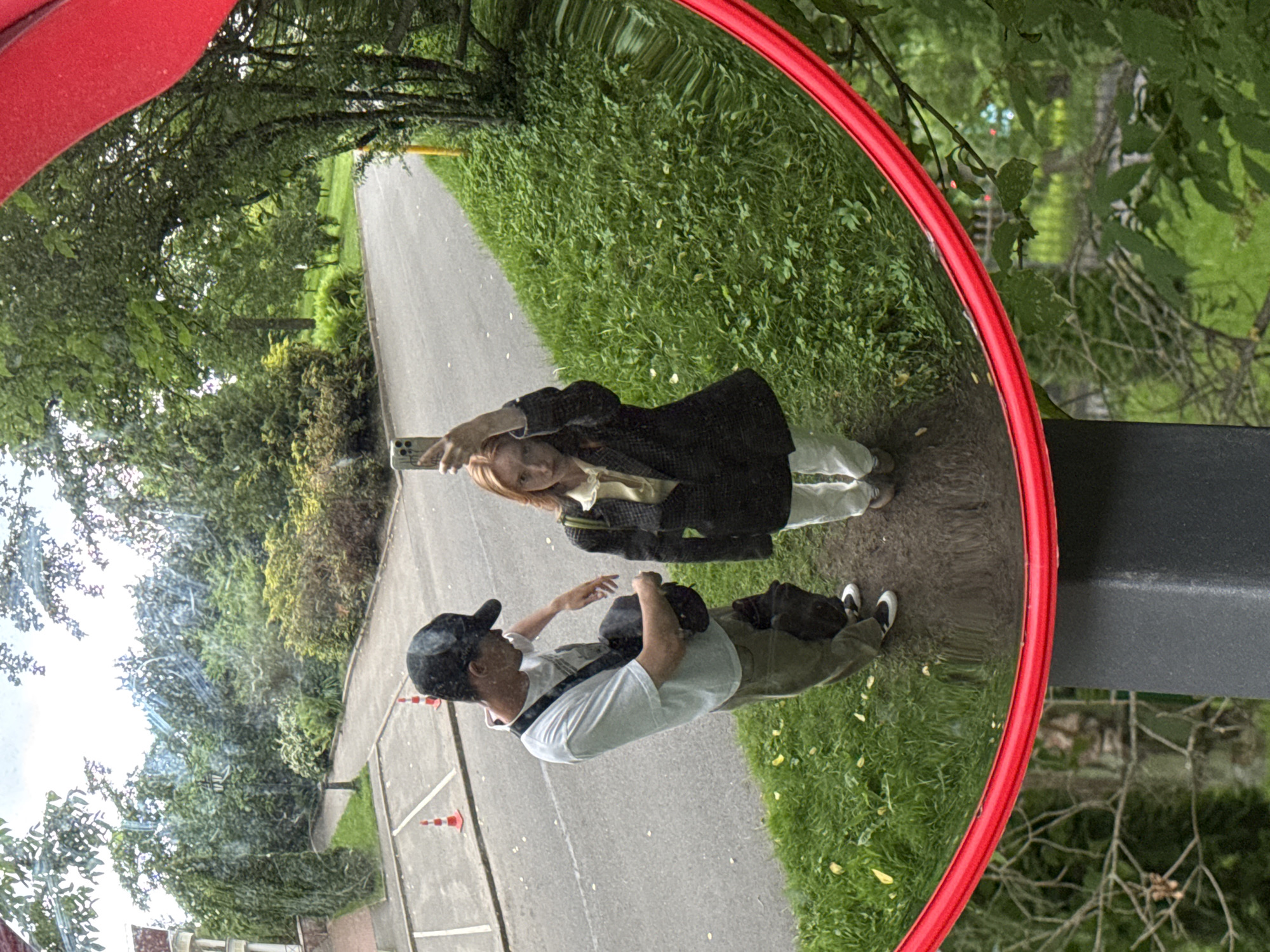 A man and a woman are reflected in a convex mirror. The man is wearing a black baseball cap, a white t-shirt, and khaki pants, and is carrying a dark-colored bag across his chest. The woman is wearing a yellow blouse, a dark plaid blazer, and white pants. She is holding a smartphone up to take a picture. They are standing on a dirt path next to a grassy area. In the background, a road and trees are visible. The scene appears to be an outdoor public space, possibly a park or a garden, in Saint Petersburg, Russia. The mirror is framed by a bright red band and is mounted on a dark gray rectangular post.