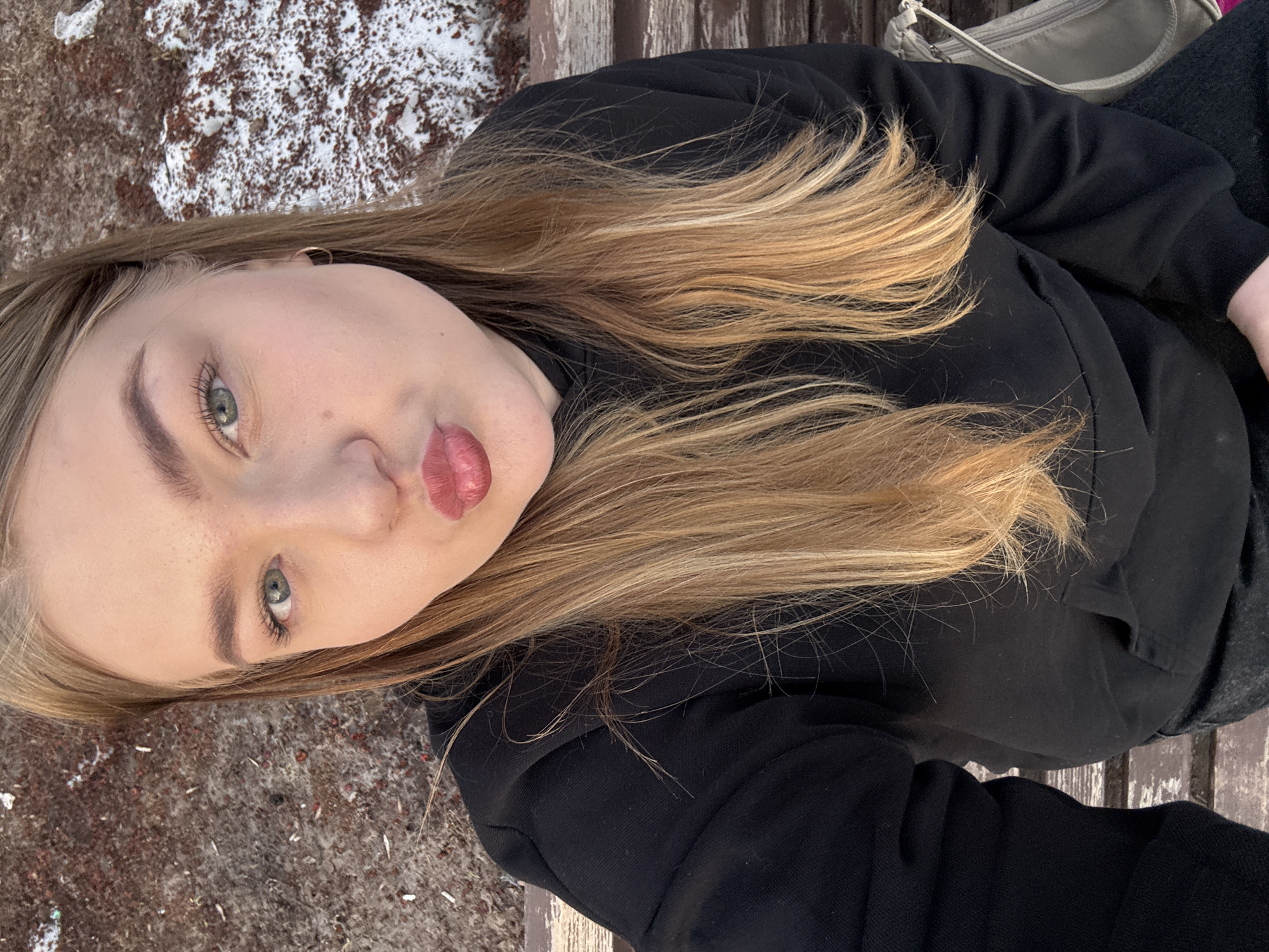A woman with blond hair and green eyes is sitting on a wooden bench, likely outdoors, based on the surrounding background. The woman is wearing a black sweatshirt and dark pants. A gray purse is visible next to her on the bench. The ground around the bench shows signs of snow and debris.
The location is likely in the city of Chaikovsky, Russia.