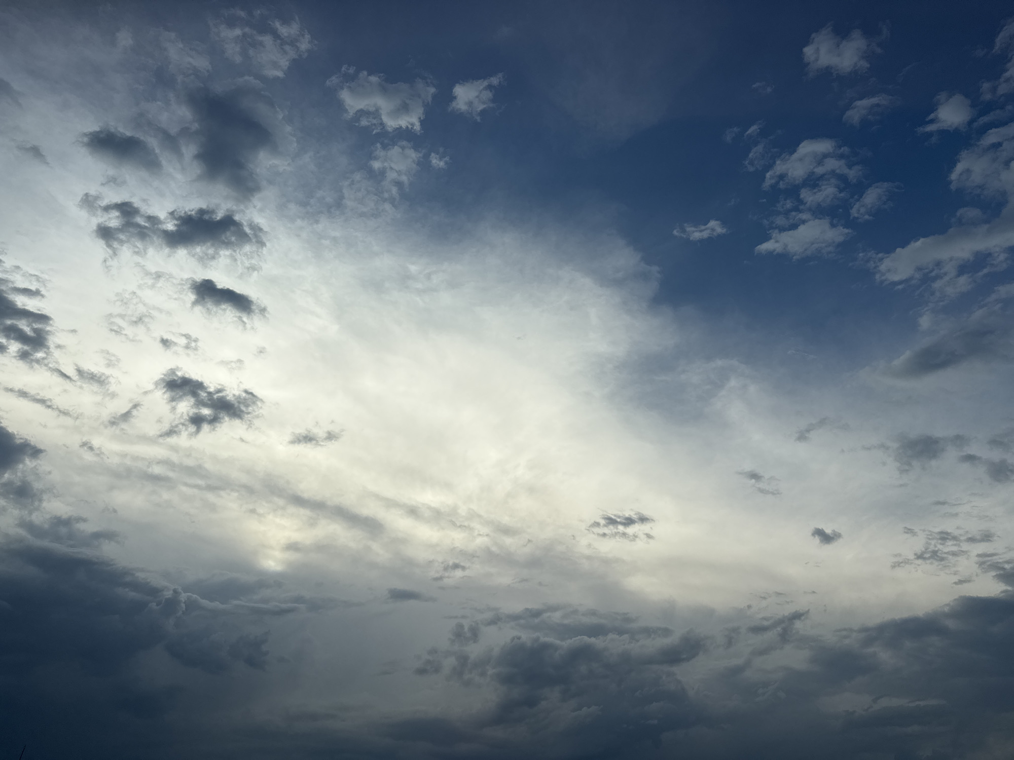 The image displays a wide, atmospheric view of a cloudy sky over Sofia, Bulgaria. The scene is dominated by various cloud formations and a gradient of atmospheric colors. In the upper right quadrant, patches of clear, deep blue sky are visible, dotted with small, wispy white clouds. As the eye moves towards the center and left, the sky becomes increasingly covered by clouds.
A bright, luminous band of white and light grey clouds stretches horizontally across the mid-section, suggesting strong illumination from a light source, likely the sun, positioned low or behind these clouds. Below this brighter area and towards the bottom-left of the frame, the clouds grow significantly darker and denser, appearing in deep shades of grey, blue-grey, and almost black. These heavier, more dramatic cloud masses create a strong contrast with the brighter portions of the sky.
The overall impression is one of dynamic weather conditions, possibly indicating an impending change in weather or the late afternoon/early evening light filtering through a complex cloudscape. There are no discernible people, man-made objects, or text visible in the photograph. The image captures a moment of natural beauty and the vastness of the sky.
