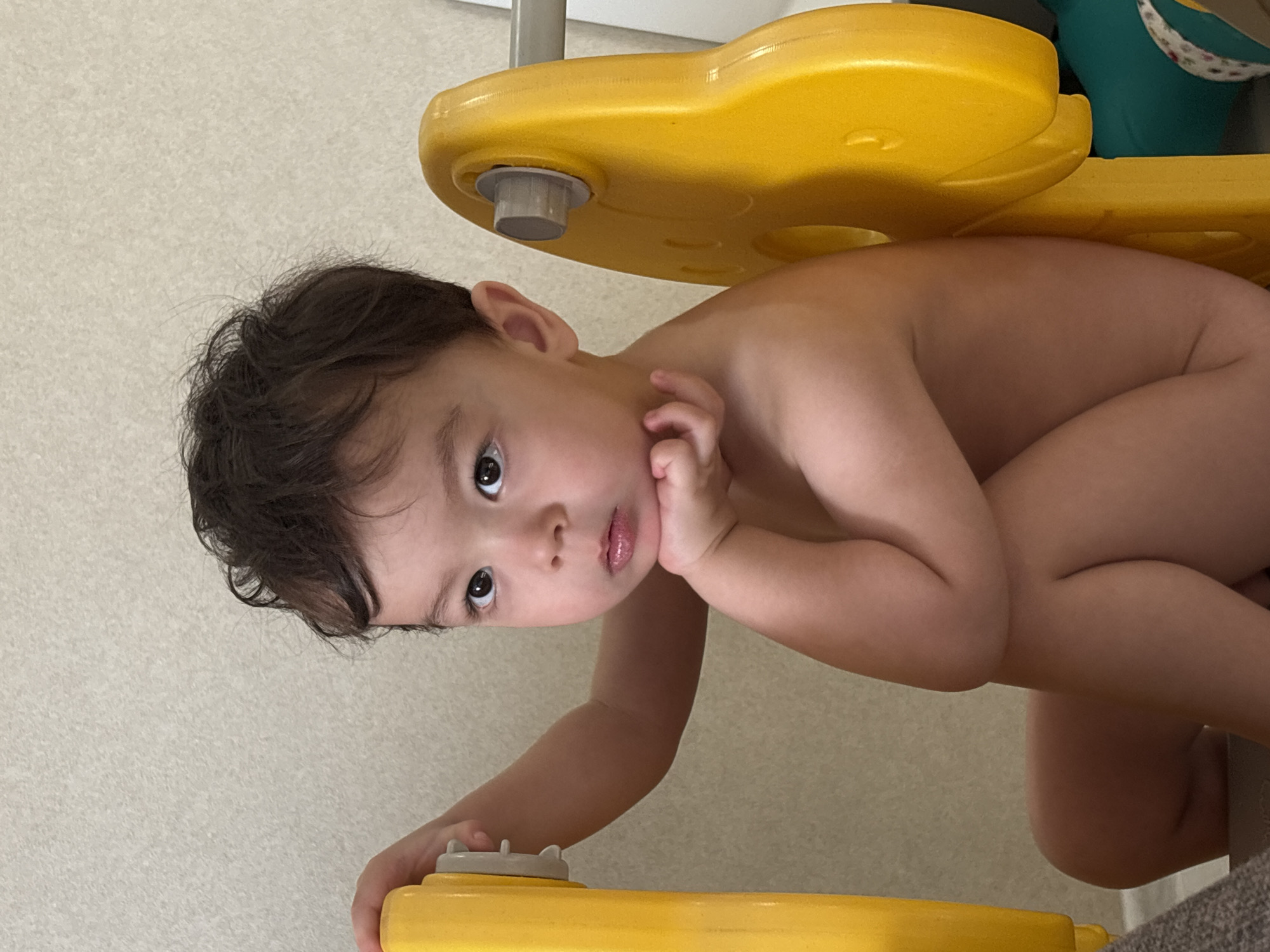 A young child with brown hair is positioned in the frame, seemingly interacting with a yellow toy. The child's right arm is extended, reaching towards the toy while their left hand is near their face. The background consists of a plain, textured wall. The location and city information are not discernible from the image.