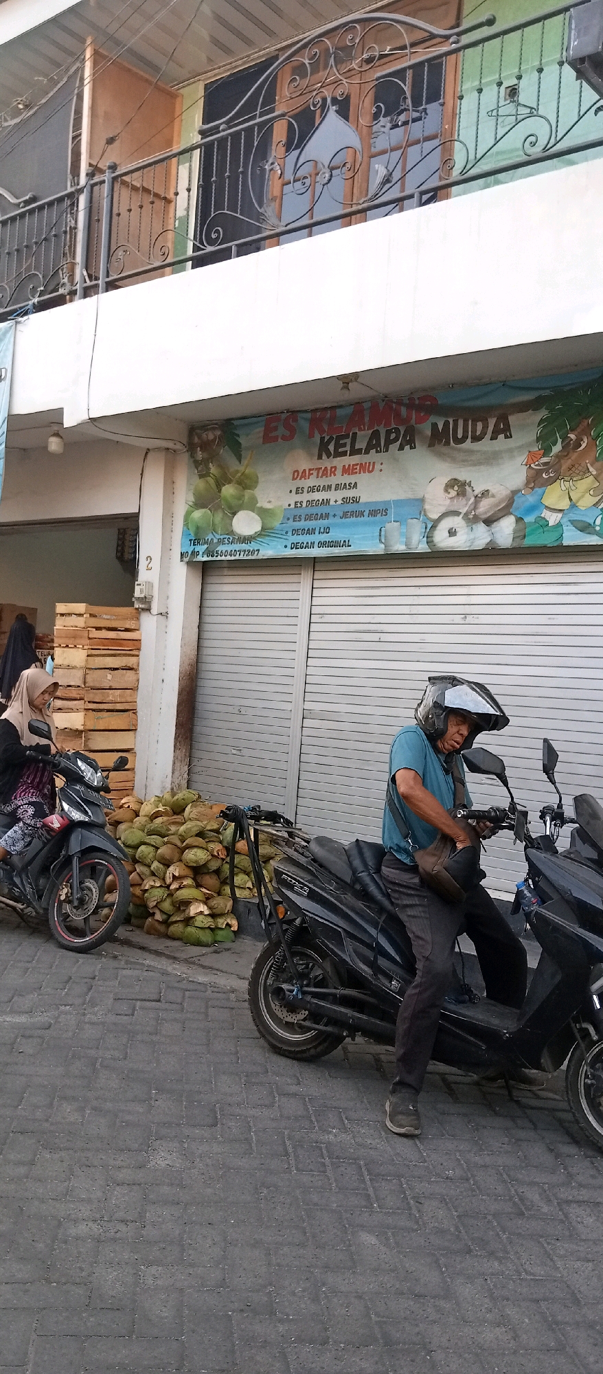 In Wadungasri, Indonesia, a street scene unfolds with a focus on a vendor selling young coconuts. A man, wearing a blue t-shirt and dark pants, sits on a black scooter, adjusting a brown bag on his lap. He wears a black helmet with a clear visor. In front of him, a large pile of green young coconuts, partially husked, rests on the ground. To the left, a woman wearing a light brown hijab and patterned clothing is seated on a black scooter. Behind the coconuts and scooters, a building features a large banner advertising "ES KELAMUD KELAPA MUDA" (Young Coconut Ice), with a menu listing various coconut drinks. The building also has a metal railing above the banner and a closed metal shutter. Wooden crates are stacked to the left of the building. The ground is paved with dark interlocking bricks.